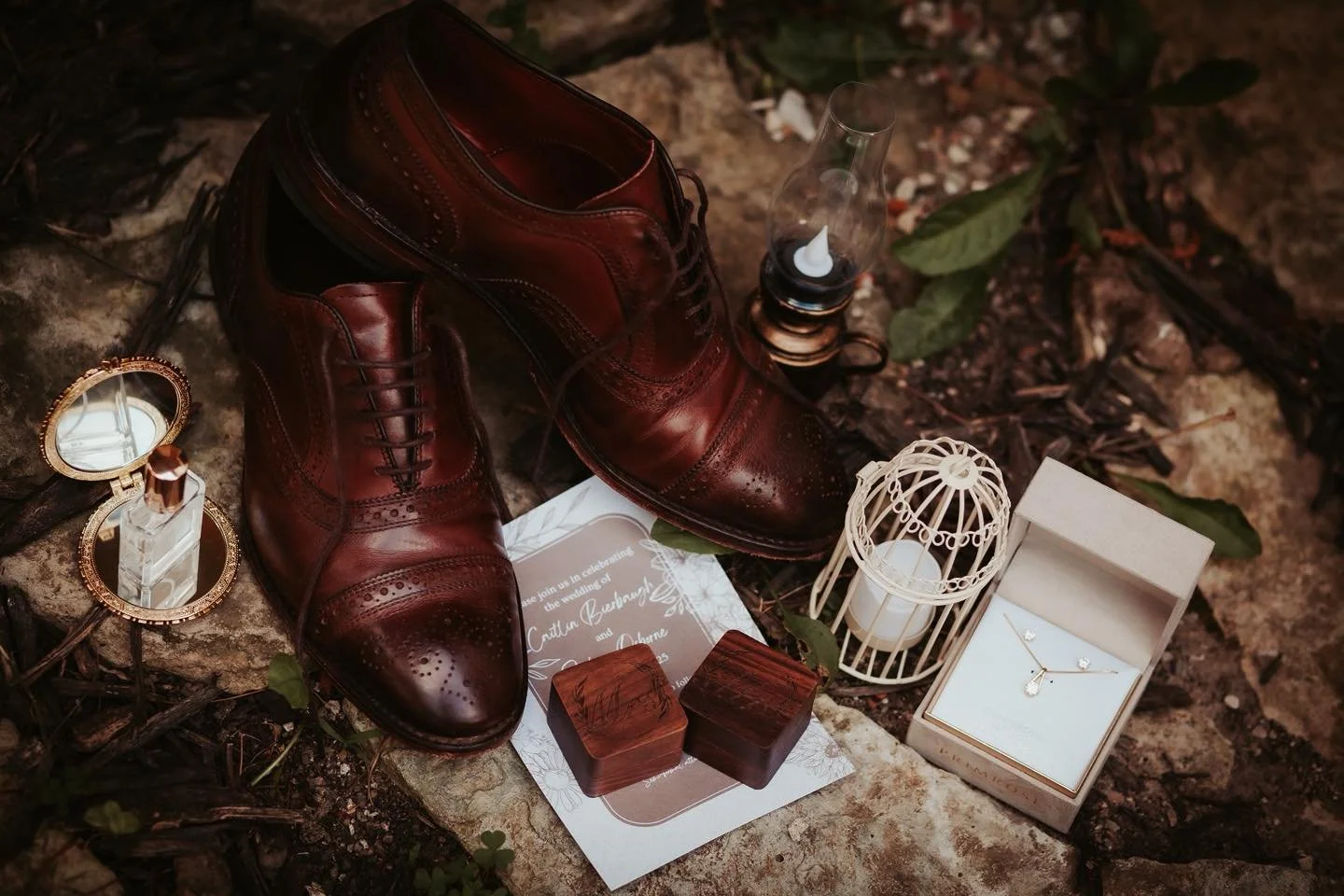 A pair of polished, dark brown leather dress shoes surrounded by jewelry, candles, decorative boxes, and green leaves on a natural outdoor surface, likely for a wedding or special event.