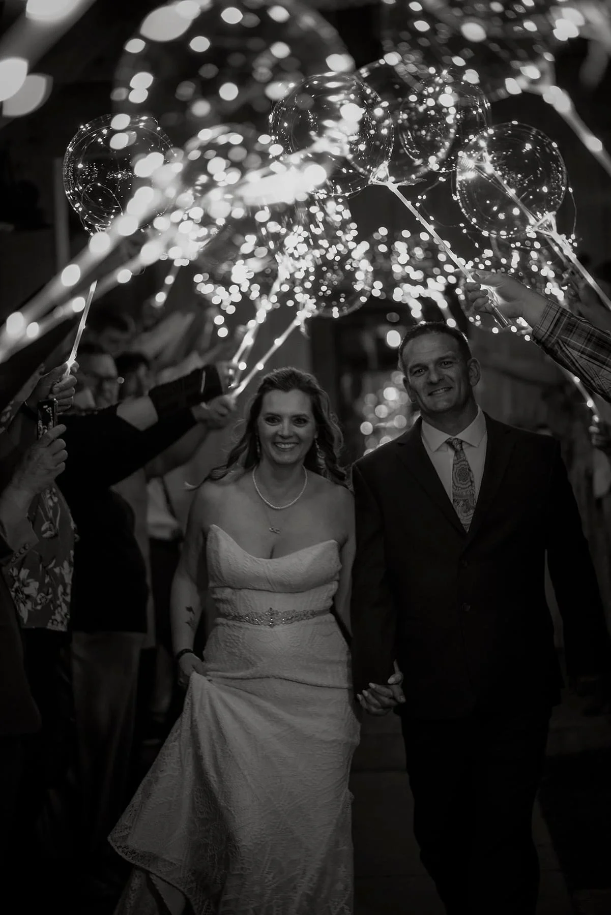 A bride and groom walking hand in hand under a tunnel of illuminated balloons, surrounded by guests celebrating at a wedding reception.