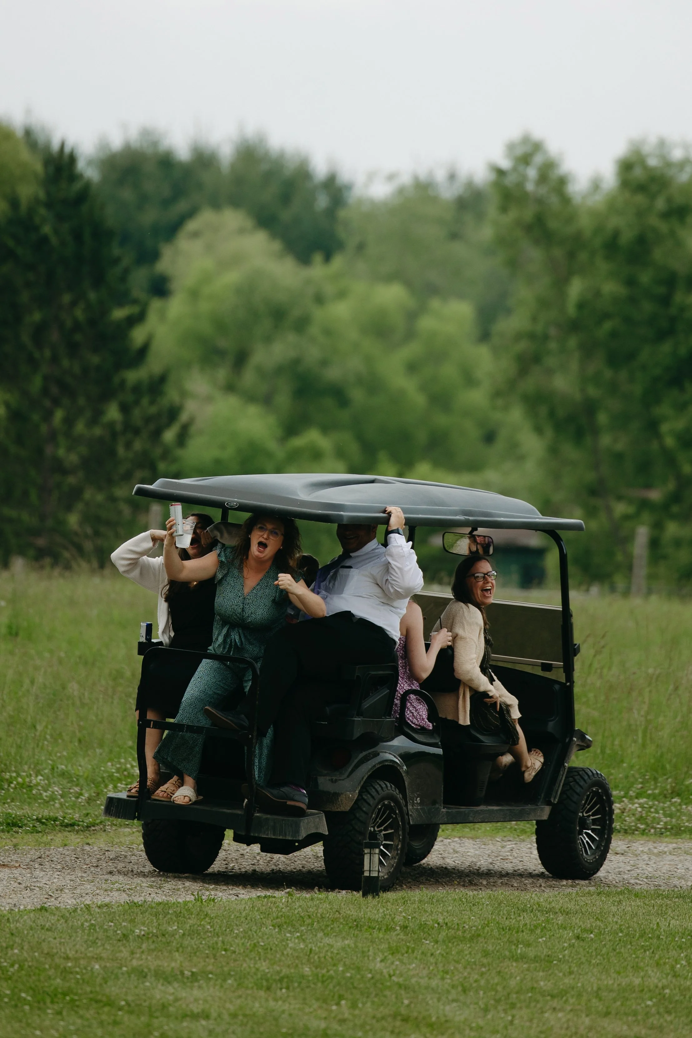 Group of five people sitting and standing on a black golf cart, smiling and laughing, in a green outdoor setting with trees and grass.