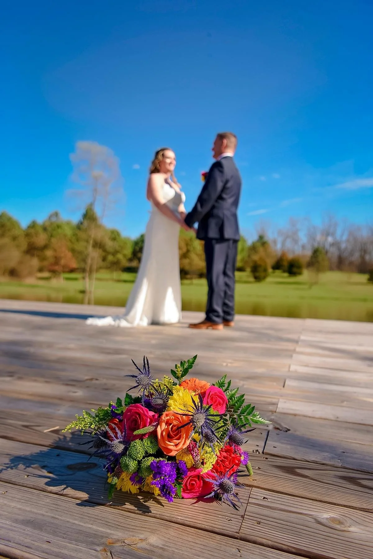Vibrant wedding bouquet with pink roses, orange flowers, yellow blossoms, and purple thistles in the foreground, with a bride and groom holding hands and smiling in the background on a wooden platform outdoors against a bright blue sky and green land