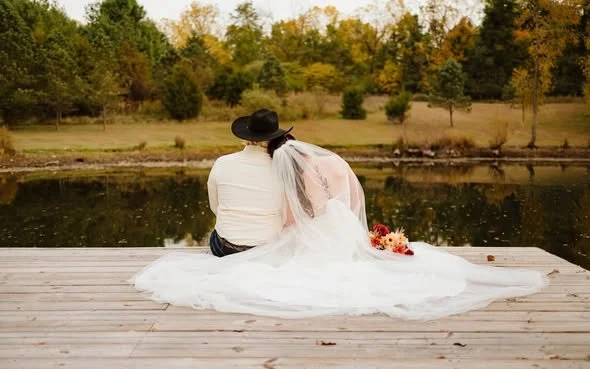 A bride and groom sitting on a wooden dock by a lake, facing away. The bride wears a white wedding dress and veil, with a bouquet beside her. The groom wears a white shirt and a black cowboy hat. Trees with fall foliage are in the background.