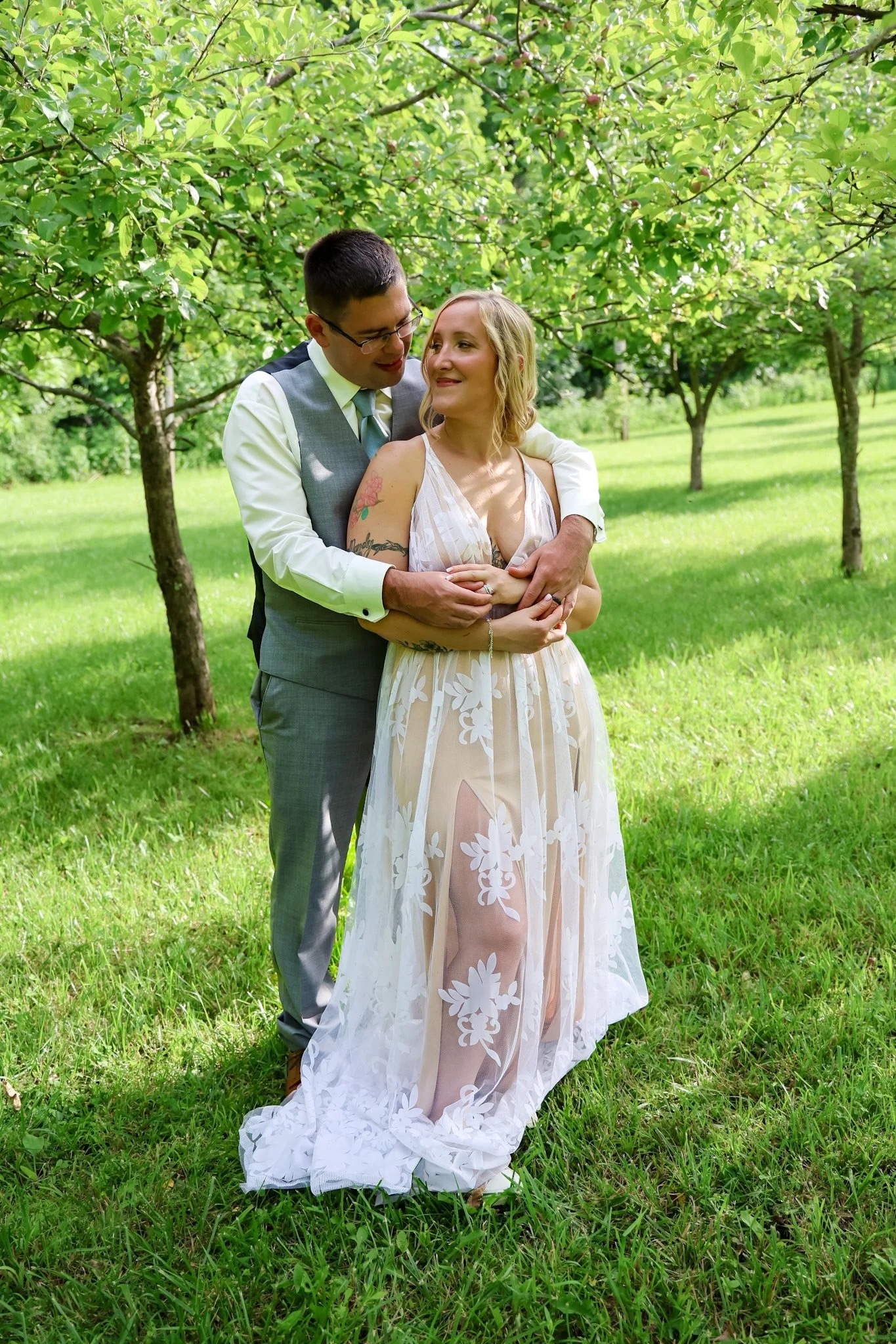 A couple stands close together in a lush green park under a tree, embracing and looking at each other, dressed in wedding attire, with the woman in a white, semi-sheer dress with floral patterns and the man in a gray vest and pants.