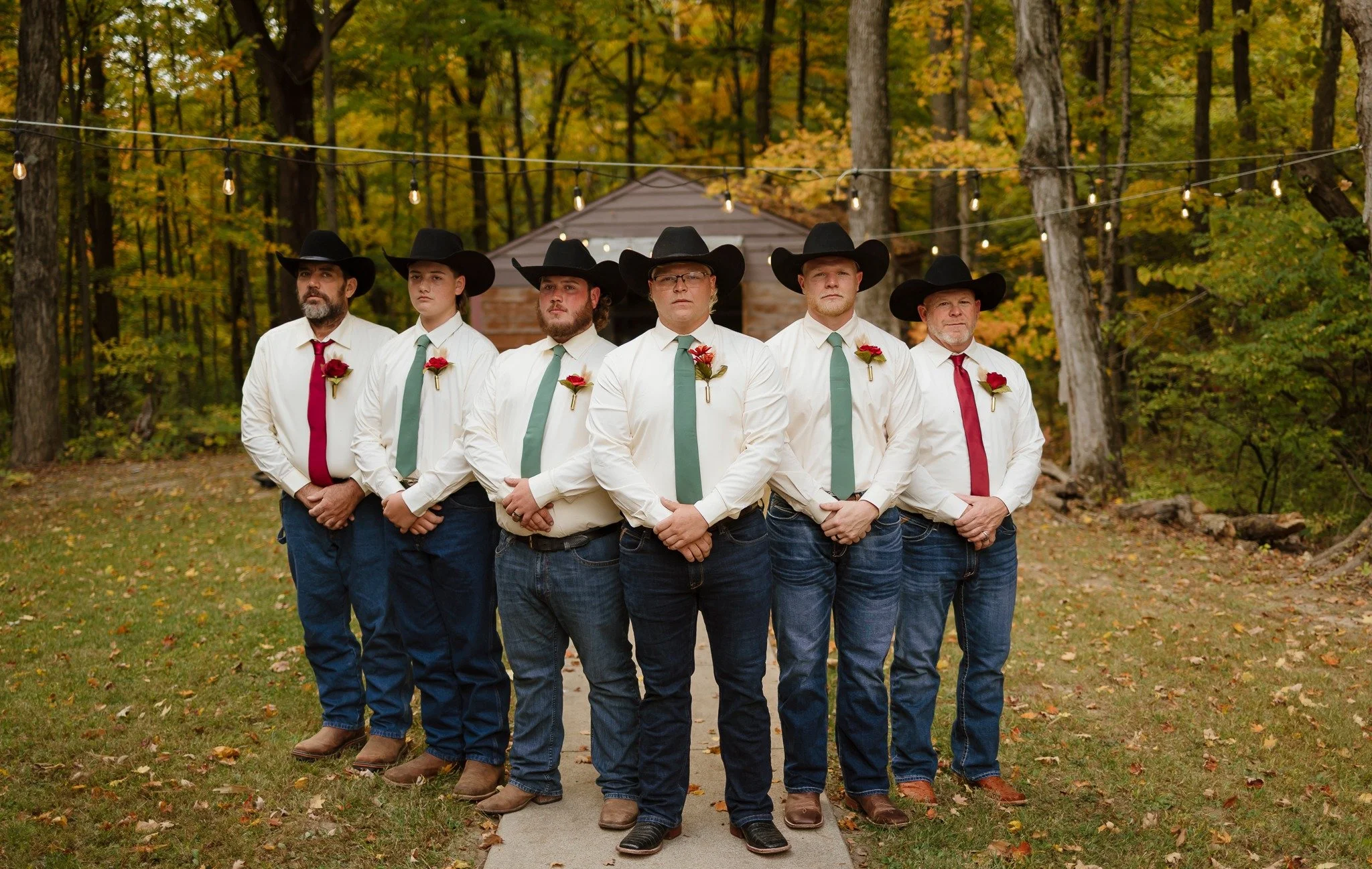 A group of six men standing in a line outdoors during fall, dressed in white shirts, jeans, cowboy hats, and ties with roses pinned to their shirts, with string lights overhead and a wooded background.