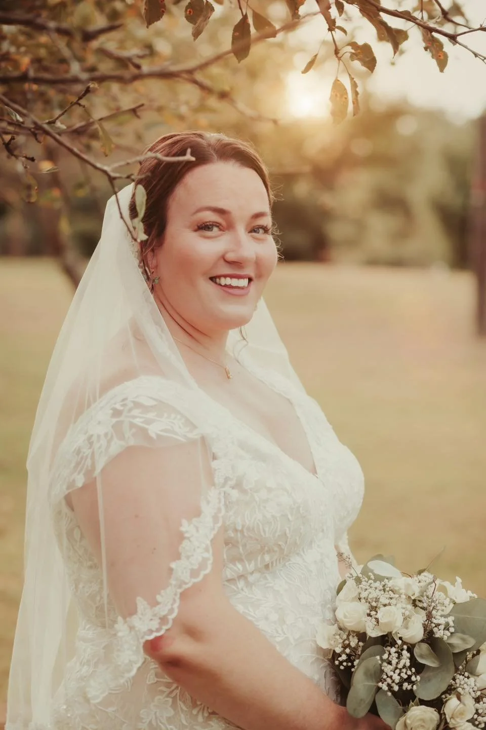 A smiling bride in a white lace wedding dress with a veil, holding a bouquet of white roses and eucalyptus leaves, outdoors with trees and sunlight at sunset.