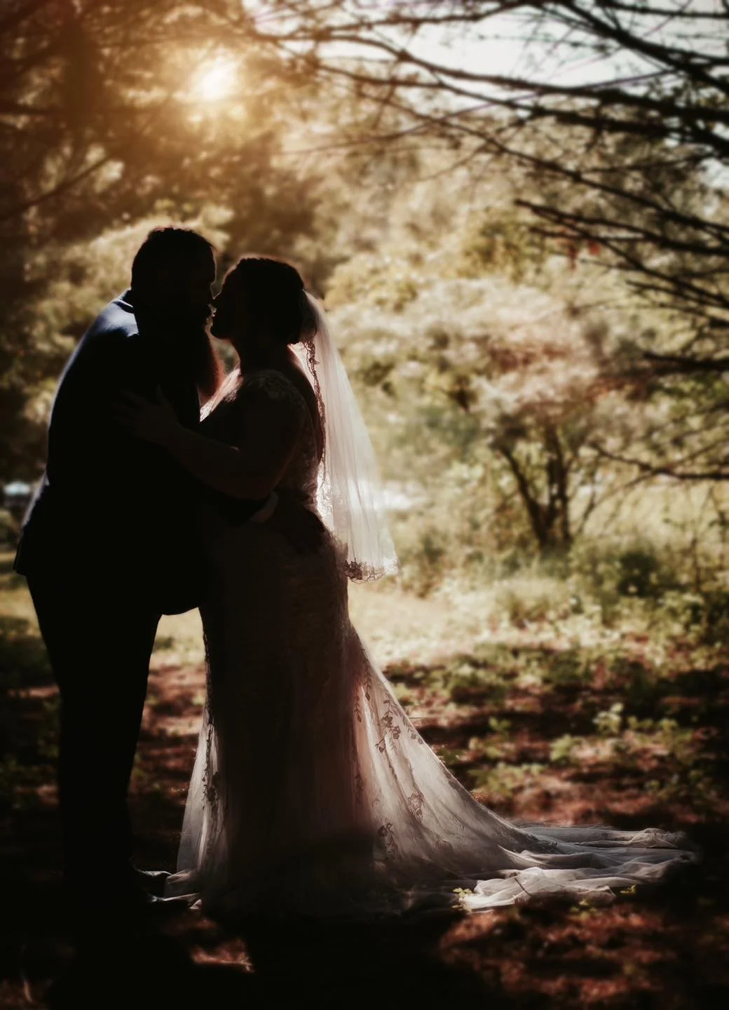 Silhouetted couple, possibly bride and groom, standing close in a wooded area during sunset, sharing a tender moment.