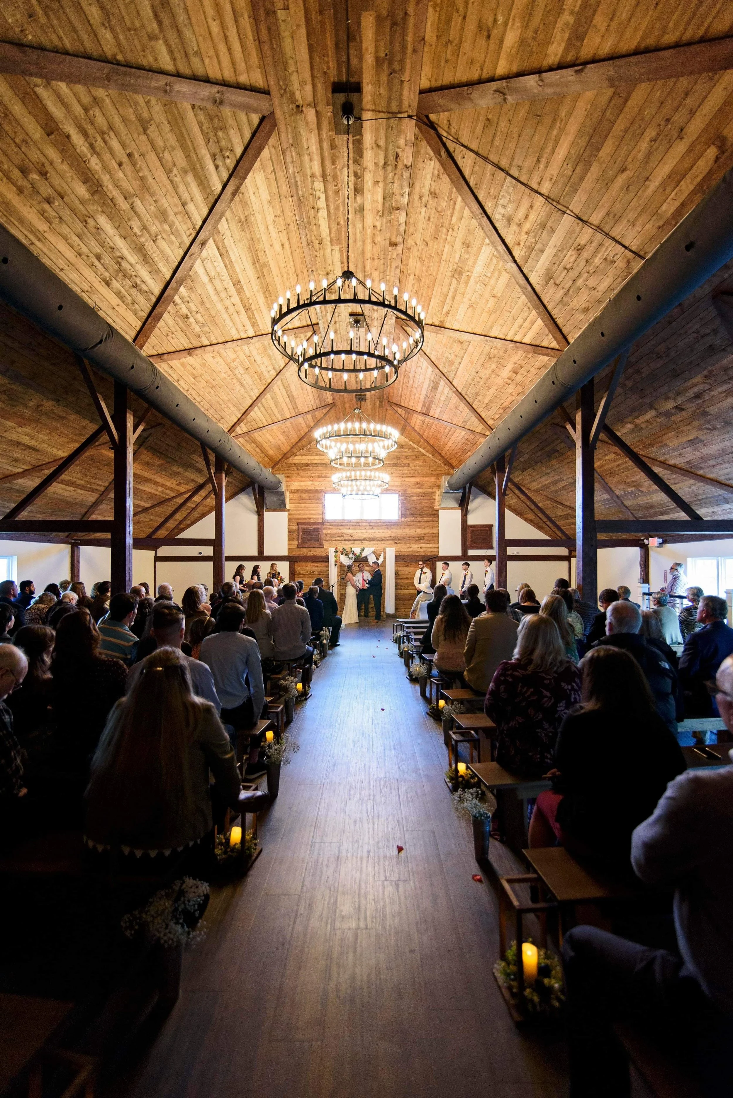 A wedding ceremony inside a rustic barn with a wooden ceiling and chandeliers. Guests are seated on benches facing the couple at the altar.