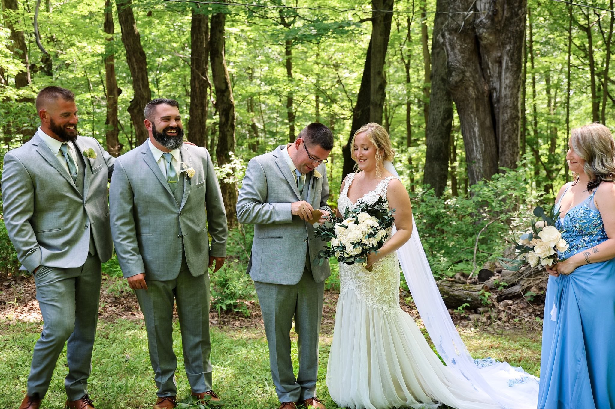 A wedding ceremony taking place in a forest with six people, including a bride in a white wedding dress holding a bouquet, and five guests in suits and dresses, standing on grass among trees.