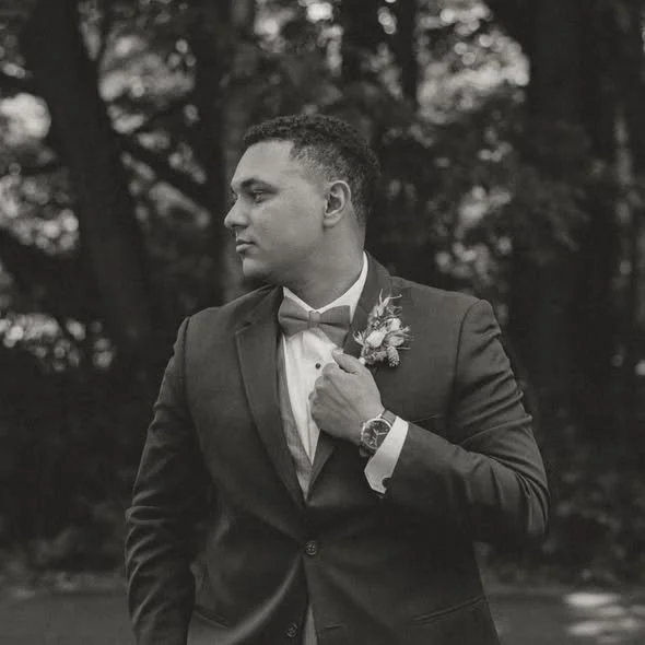 A young man in a tuxedo with a bow tie and boutonniere, standing outdoors with trees in the background, adjusting his lapel.