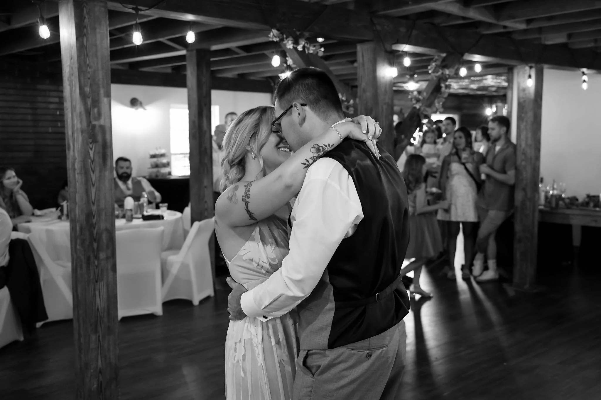 A couple is dancing and embracing at a wedding reception, surrounded by guests in a rustic venue with exposed wooden beams and string lights.