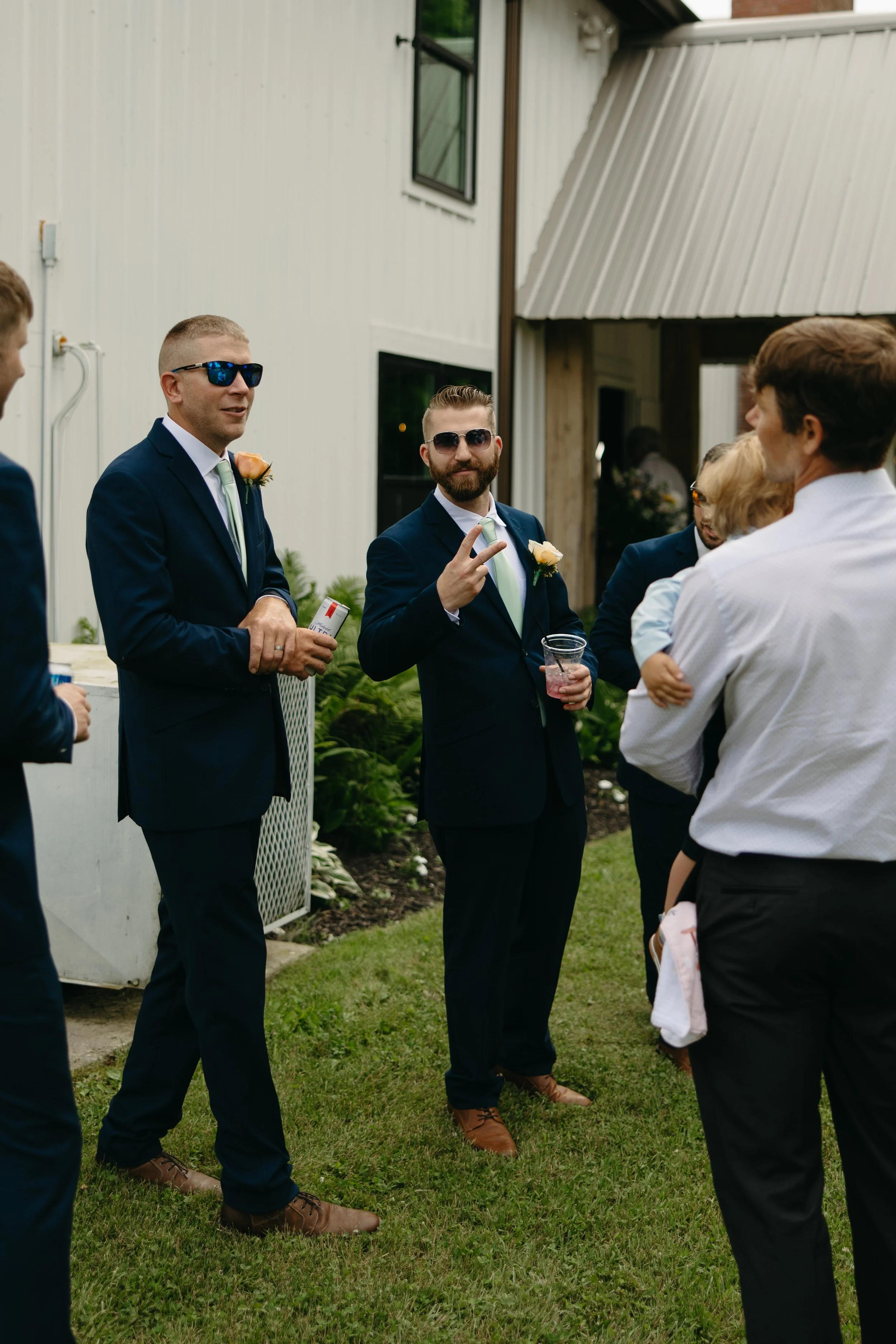 Men in suits at an outdoor gathering, one making a peace sign, in front of a white building with open windows.
