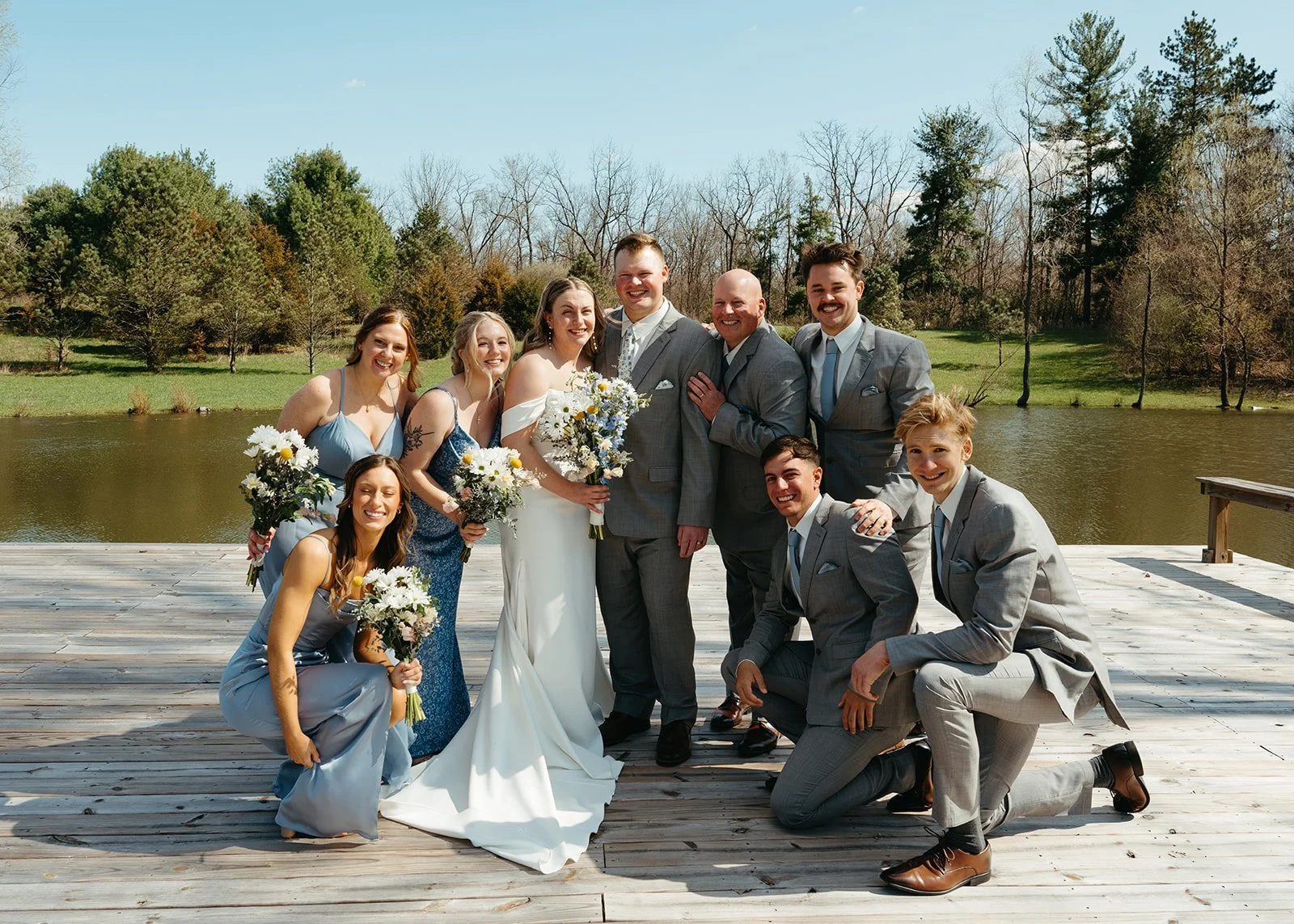 A wedding party of ten people, including the bride and groom, posing on a wooden dock by a lake surrounded by trees.