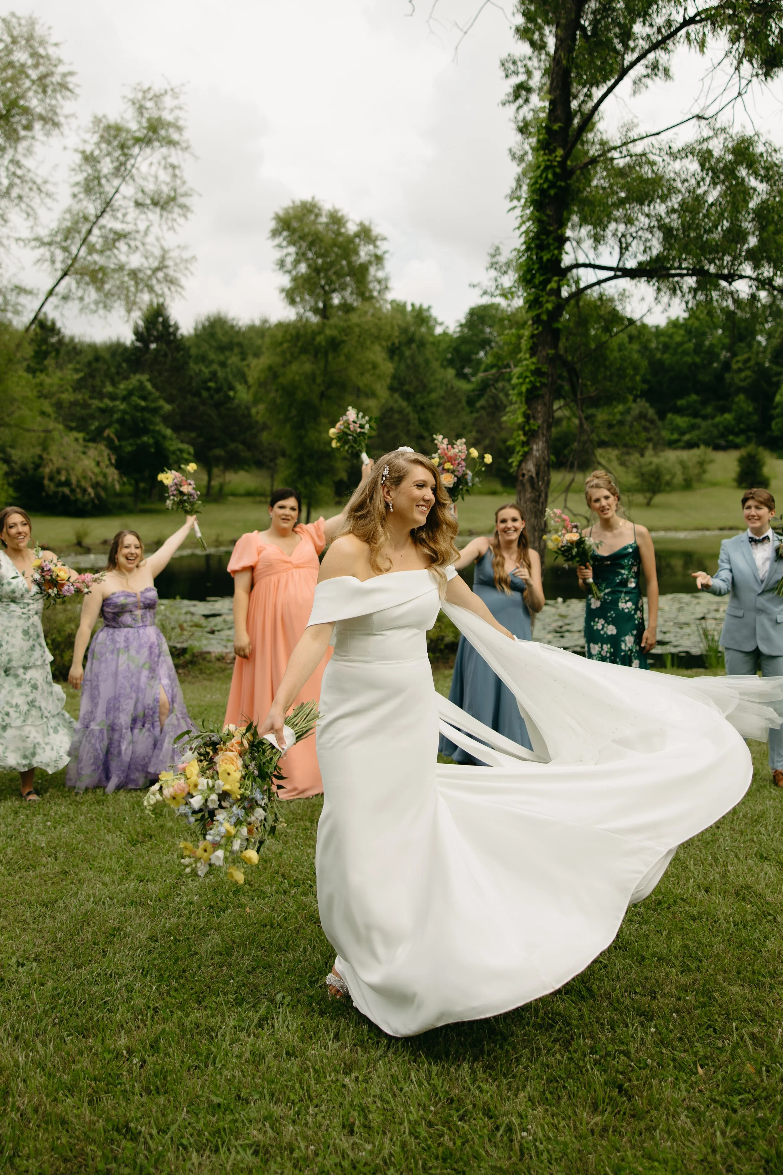 Bride walking with a flowing white dress and bouquet in front of female guests in colorful dresses by a pond.