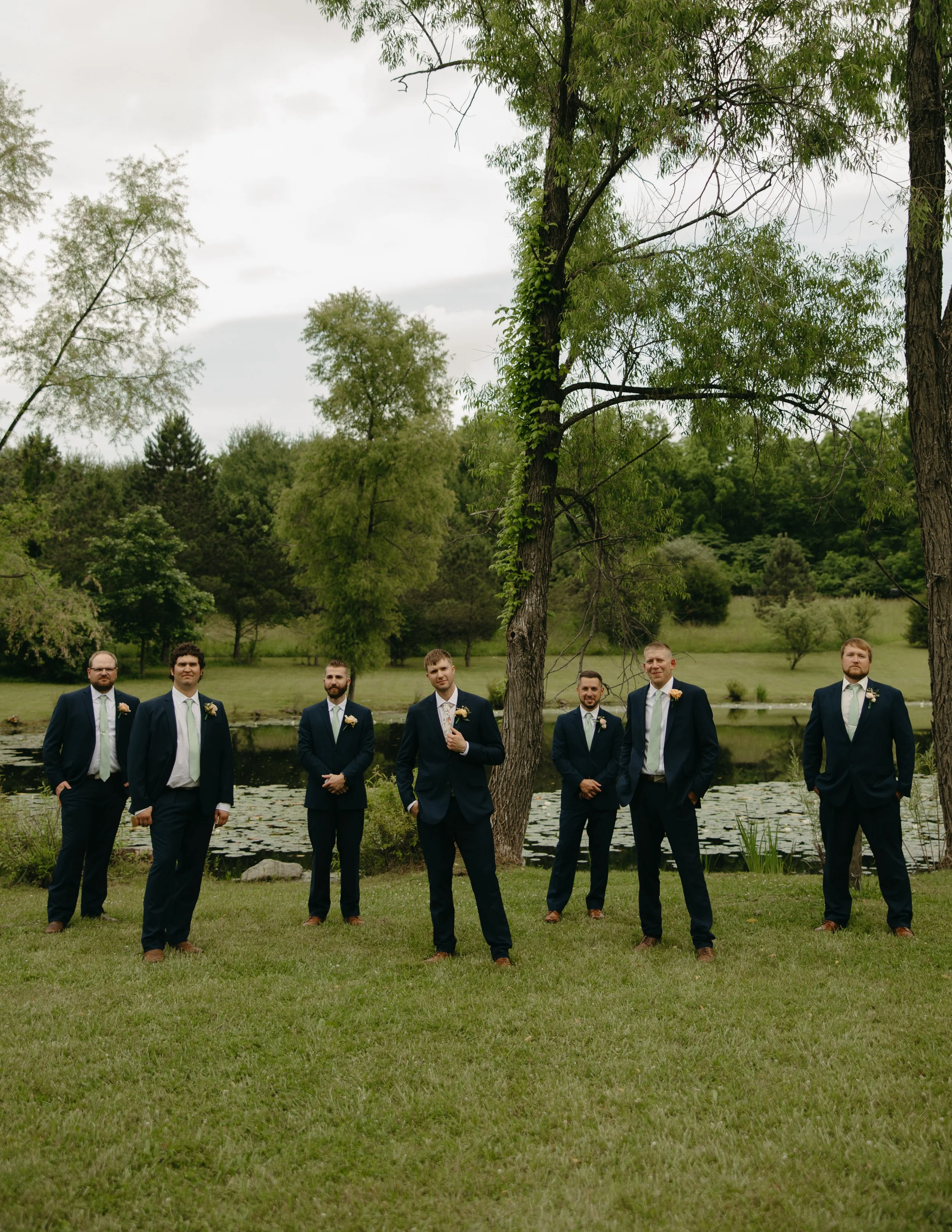 A group of seven men dressed in suits and ties standing outdoors near a pond with lily pads, surrounded by green trees, on a cloudy day taken at an Ohio wedding venue