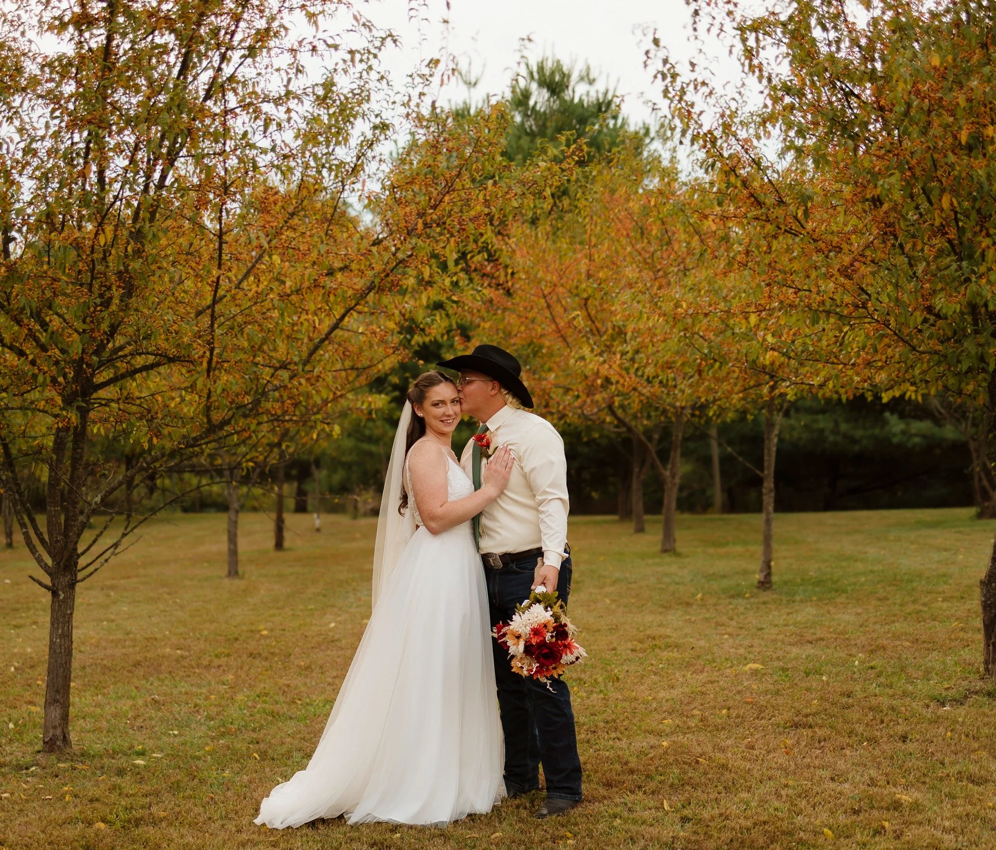 A bride and groom share a kiss outdoors during fall, with colorful autumn trees in the background.