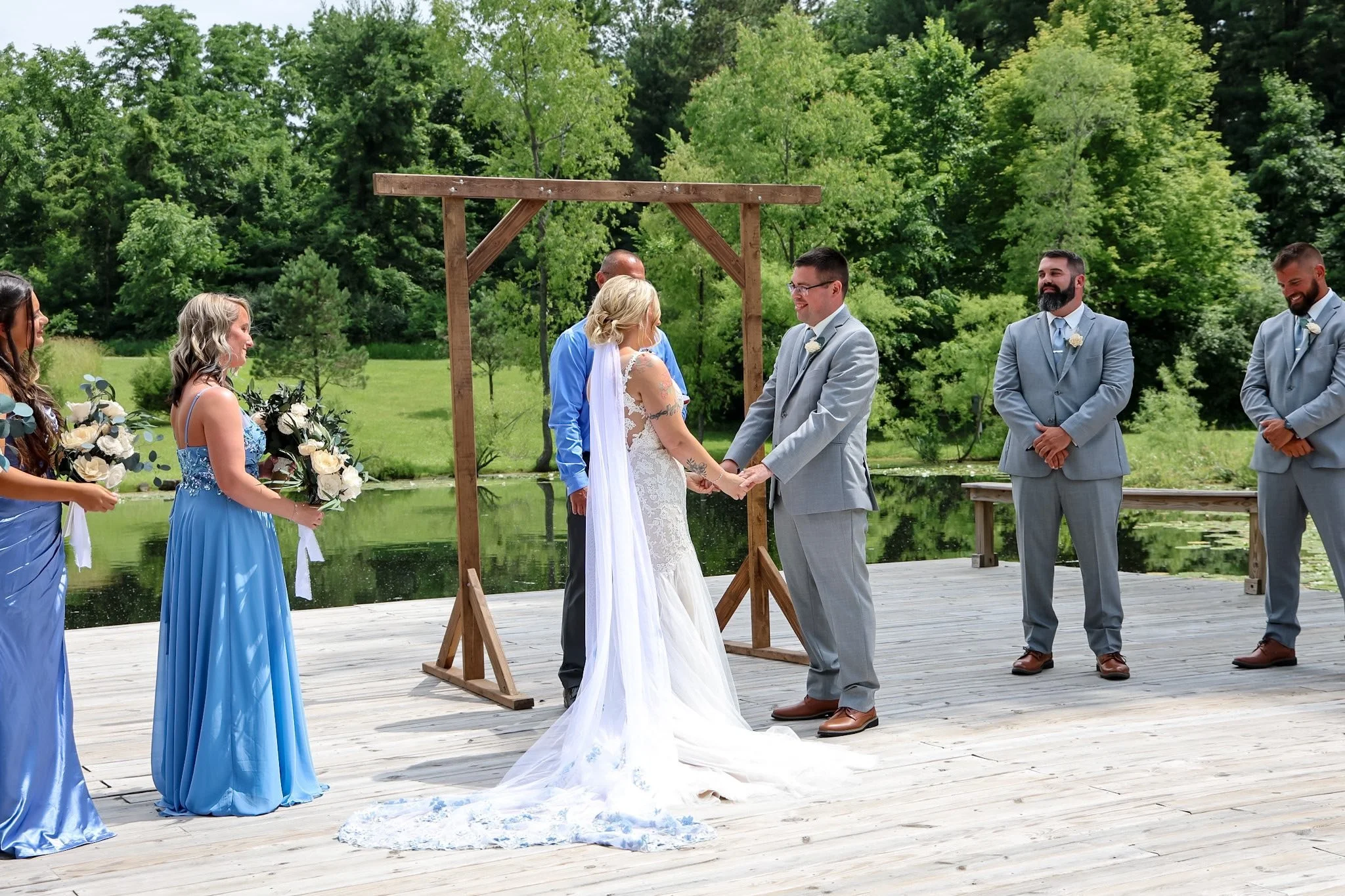 A couple getting married outdoors on a wooden platform near a pond, with friends and family present.