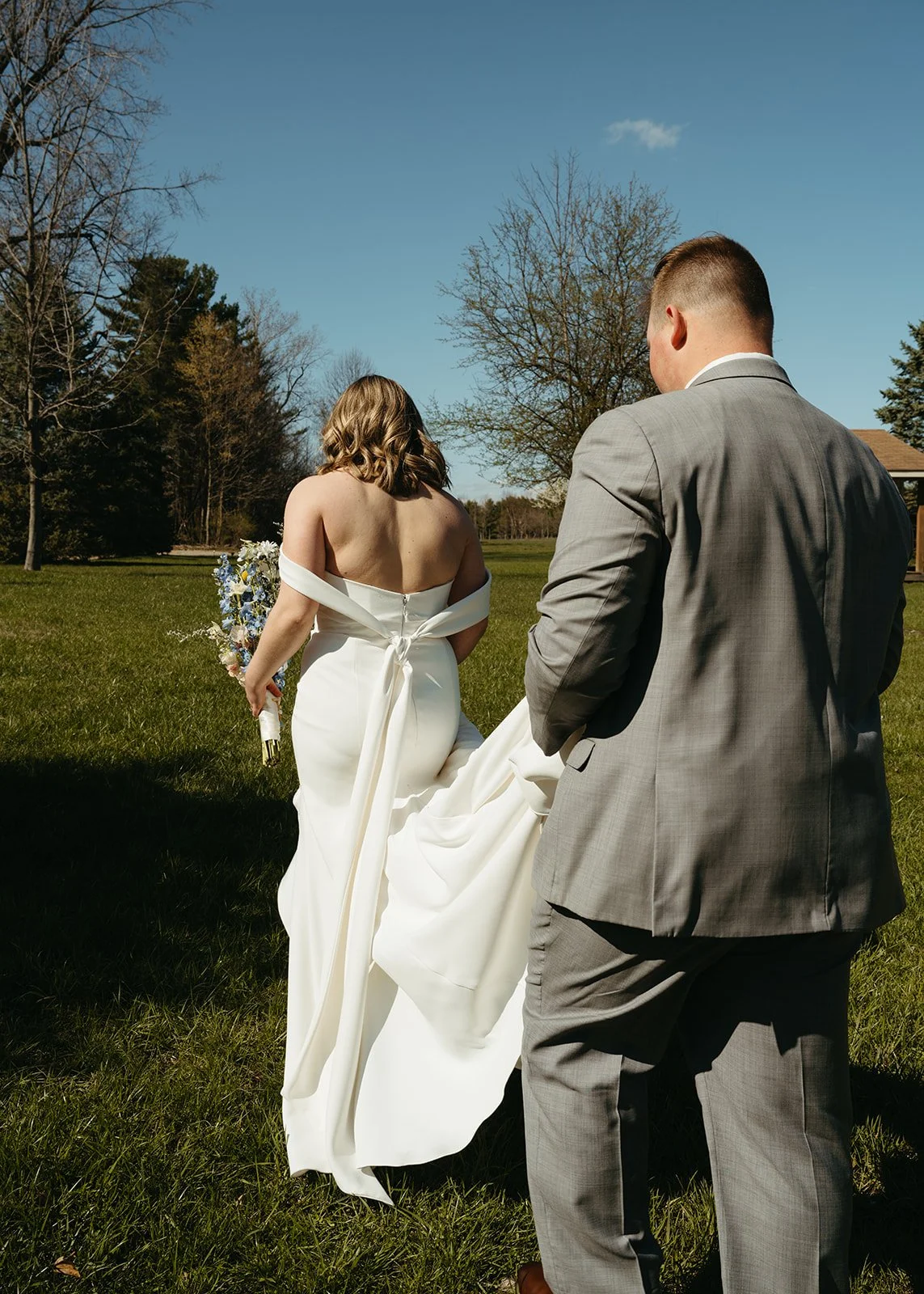 Couple in wedding attire outdoors on grassy field, woman in white gown holding bouquet, man in gray suit, trees and blue sky in background.