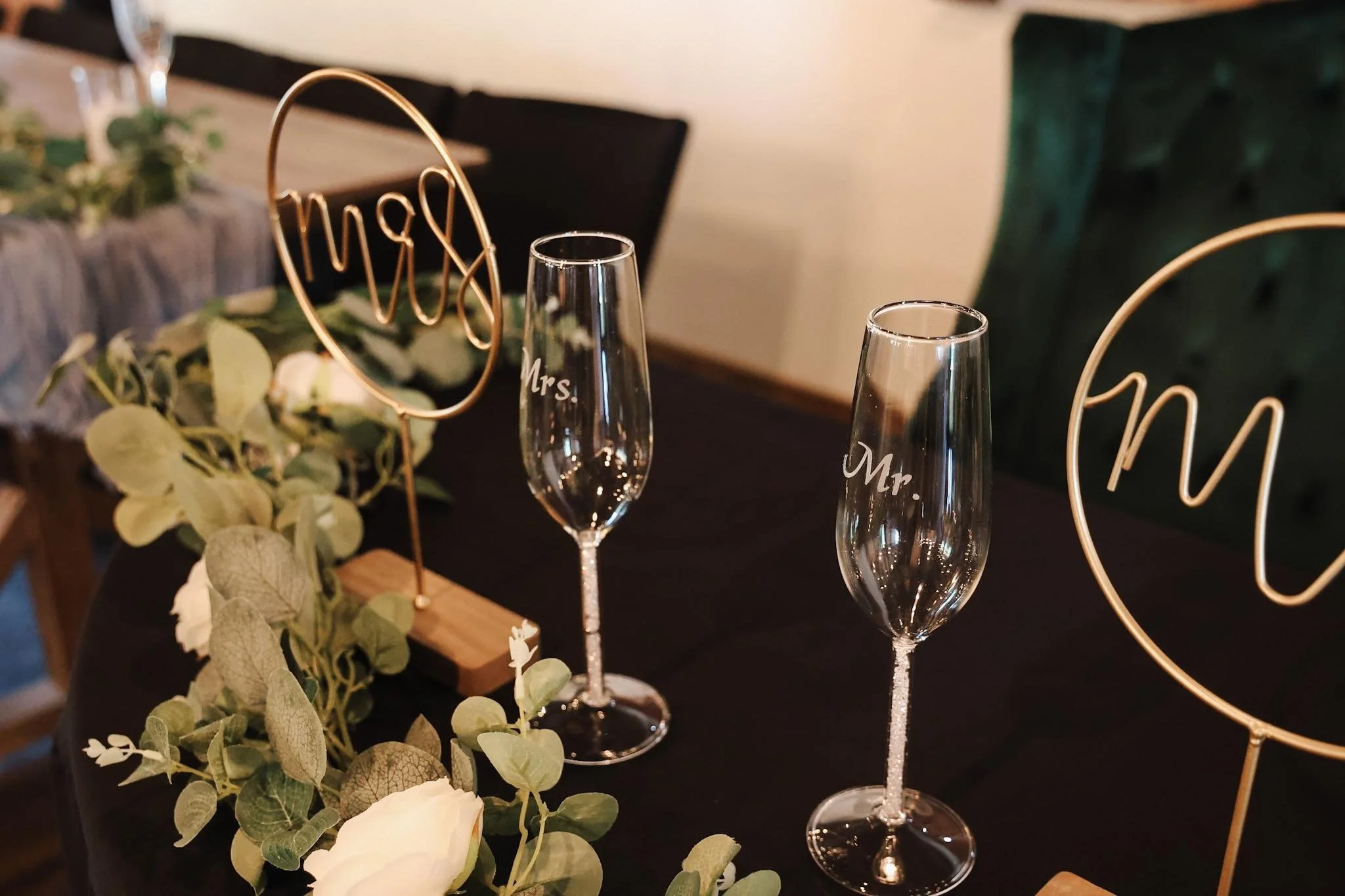 Wedding table setting with two champagne flutes labeled 'Mrs.' and 'Mr.', green foliage, white flowers, and decorative 'Mrs.' signs on a black tablecloth.