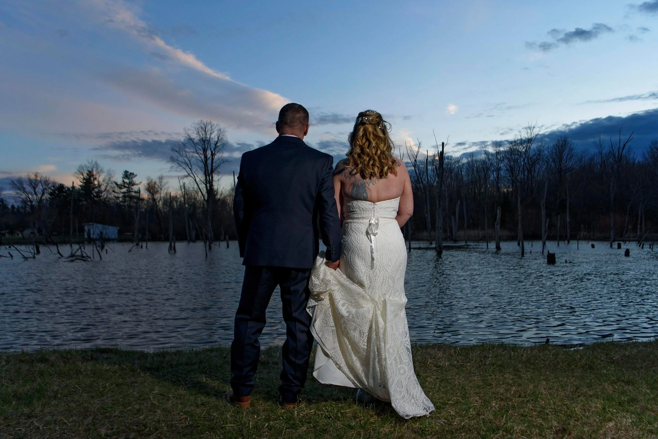 A couple in wedding attire standing by a lake at sunset, holding hands, with bare trees and a cloudy sky in the background.