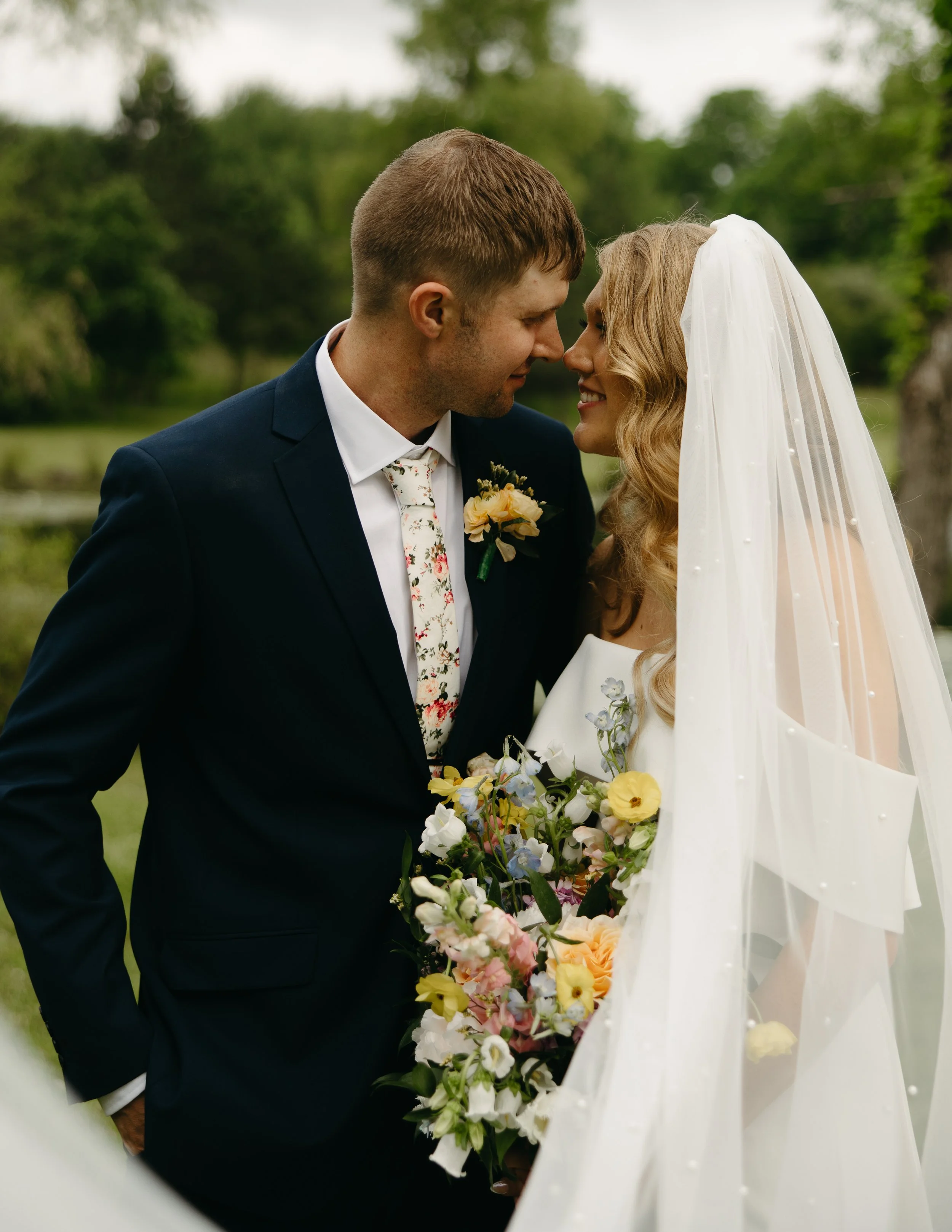 A newlywed couple, a man in a dark suit with a floral tie and a woman in a wedding dress with a veil, are standing outdoors close to each other, holding a bouquet of colorful flowers, with a scenic green landscape in the background.