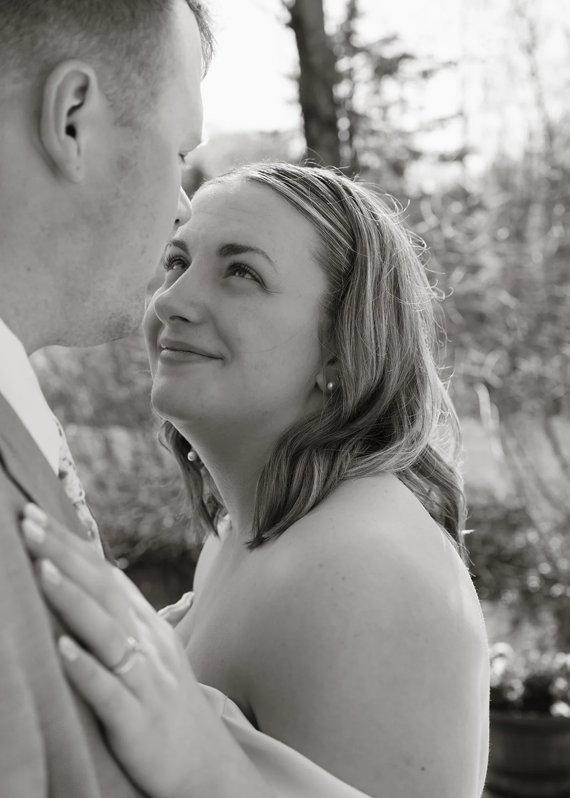 A black and white photo of a couple outdoors, gazing at each other affectionately, with the woman looking up at the man. The woman has shoulder-length hair and is wearing earrings, while the man has short hair and freckles. Tree branches are visible 