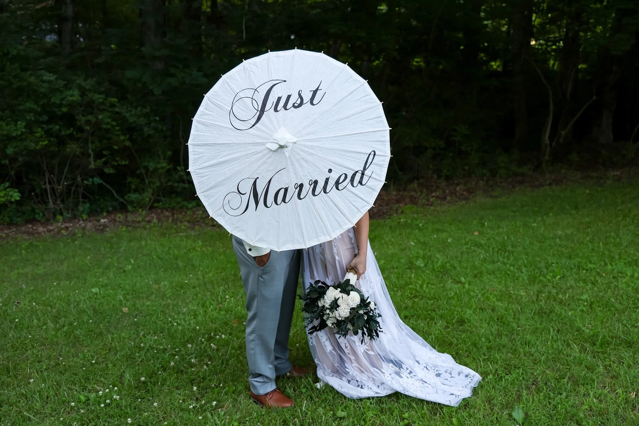 A bride and groom are standing on grass, holding a white parasol with the words "Just Married" written on it in black script. The bride is in a white wedding dress holding a bouquet of white flowers, while the groom is in a light gray suit. The backg