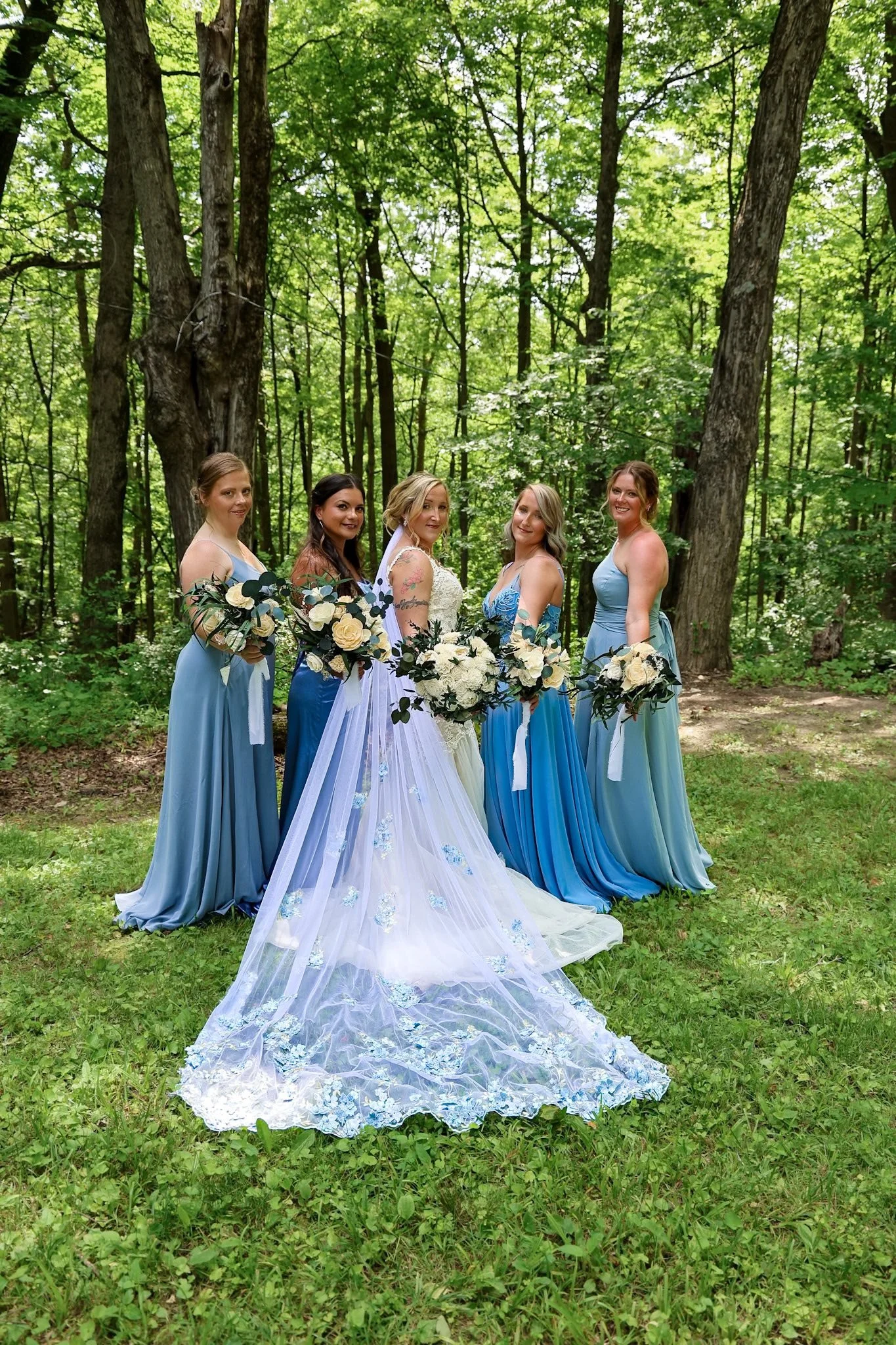 A group of five women, including a bride, standing outdoors in a forest setting with green foliage and tall trees in the background. The women are dressed in various shades of blue and are holding bouquets of white flowers. The bride is wearing a whi