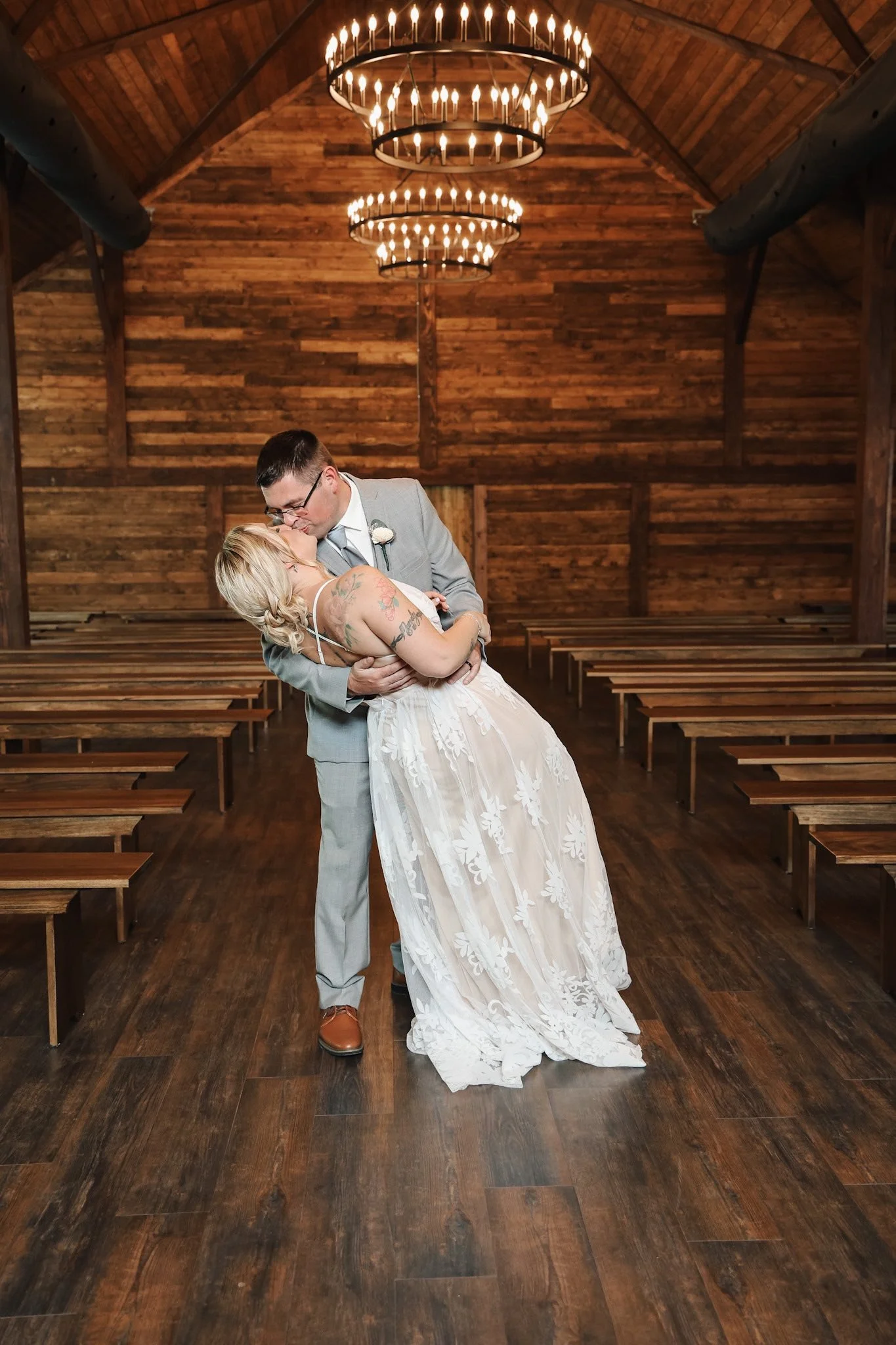 A couple kissing in a rustic wooden wedding venue. The woman wears a white wedding dress and has tattoos, while the man wears a gray suit.