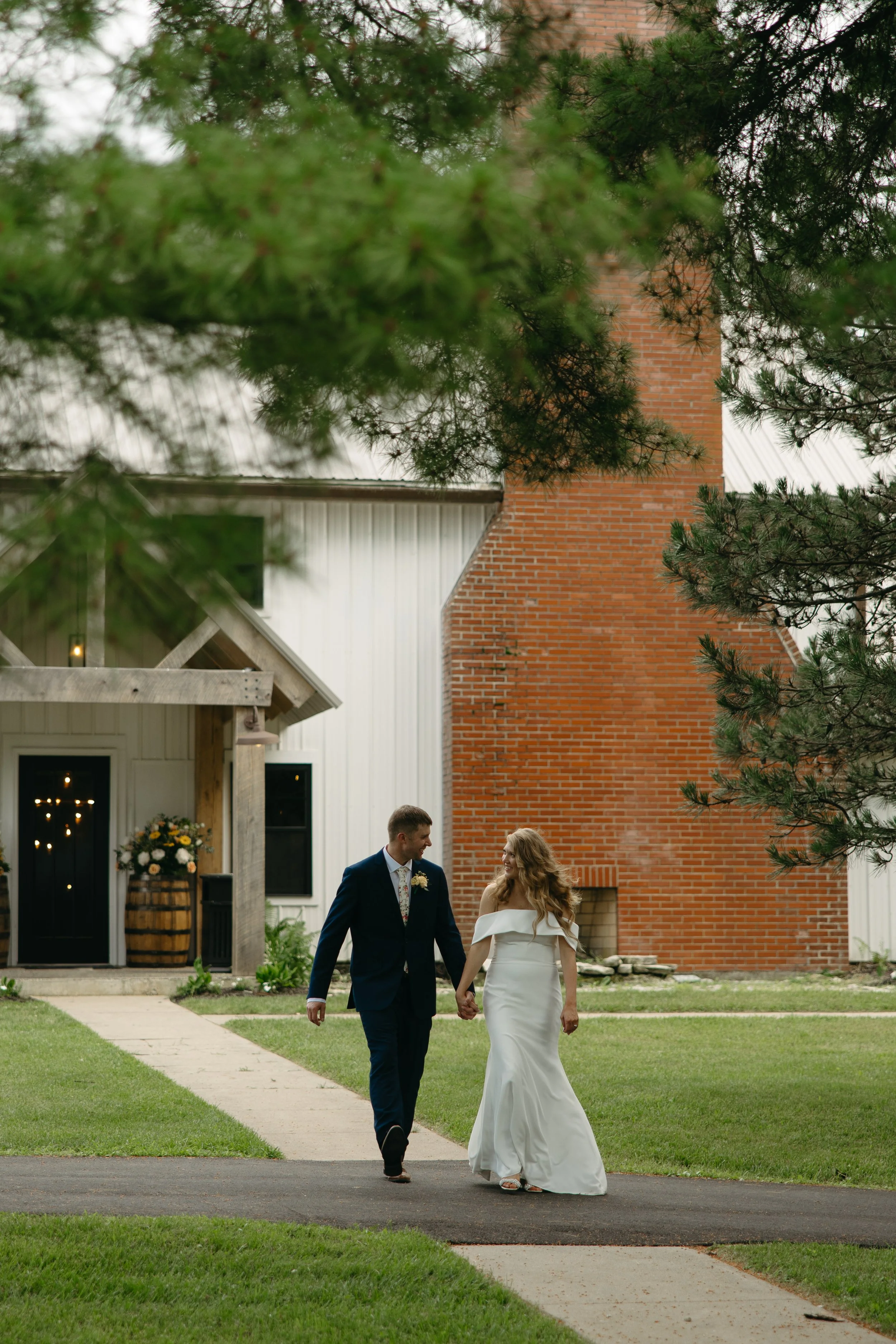 A newlywed couple walks hand in hand on a sidewalk outside a rustic wedding venue, with green grass and trees surrounding them, and a red brick chimney visible behind the venue building.