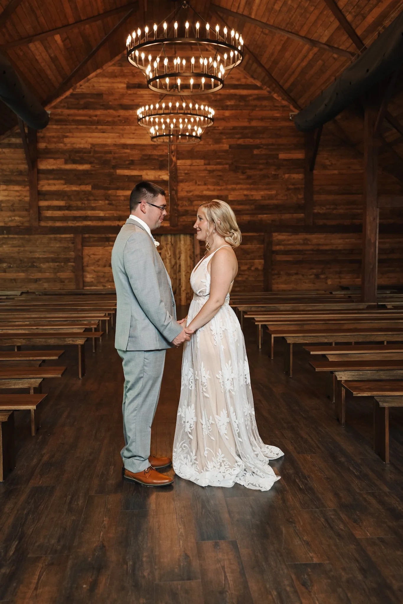 A bride and groom standing inside a rustic wooden chapel, holding hands and facing each other, with chandeliers hanging from the ceiling.