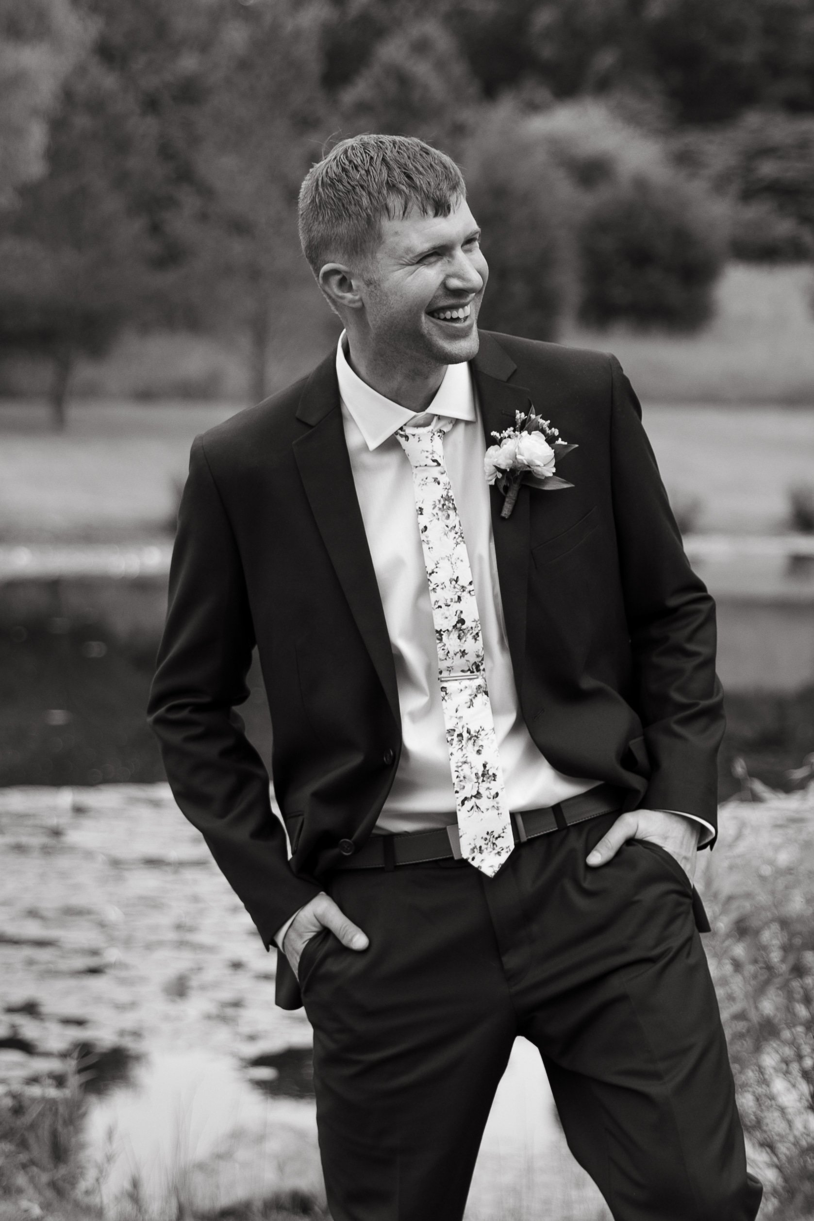 A smiling man in a suit with his hands in his pockets, standing outdoors near a small body of water.