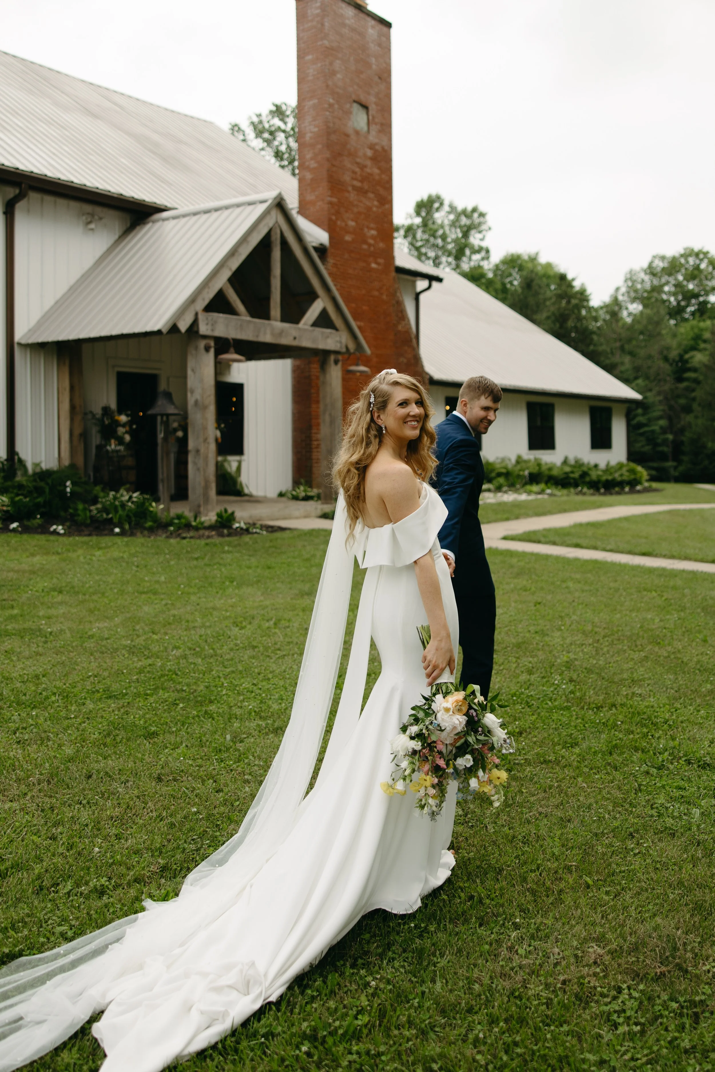 A bride in a white wedding dress with a long train holding a bouquet of flowers, standing on a grassy lawn outside a rustic building, with a smiling groom in a dark suit behind her.