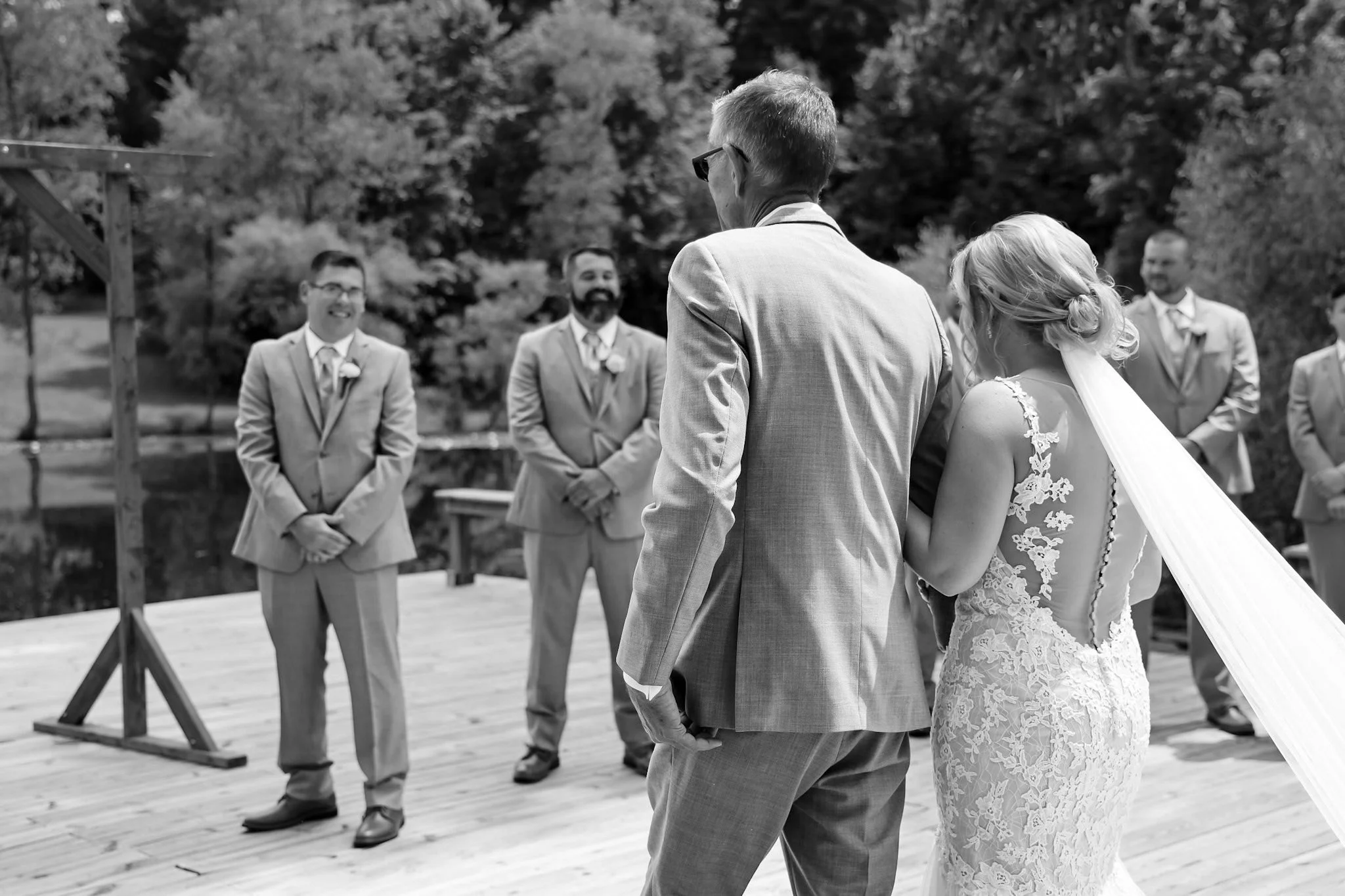 A black and white photo of a wedding ceremony outdoors with a bride and groom, surrounded by officiants and groomsmen, in a scenic area with trees in the background.