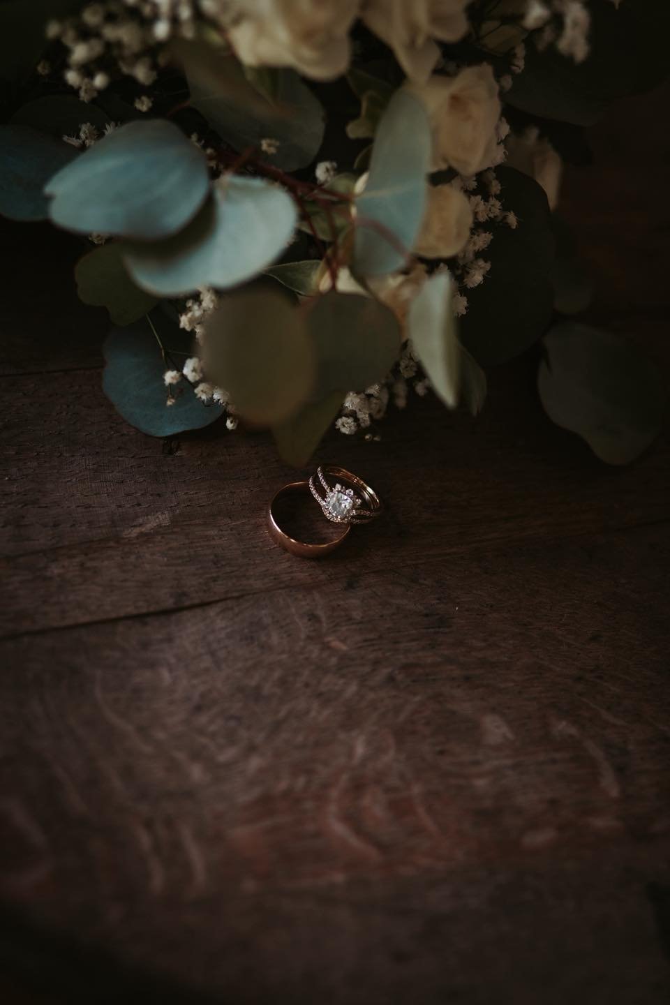 Wedding rings placed on a dark wooden surface, with a floral arrangement of green leaves, white flowers, and baby's breath partially obscuring the rings.