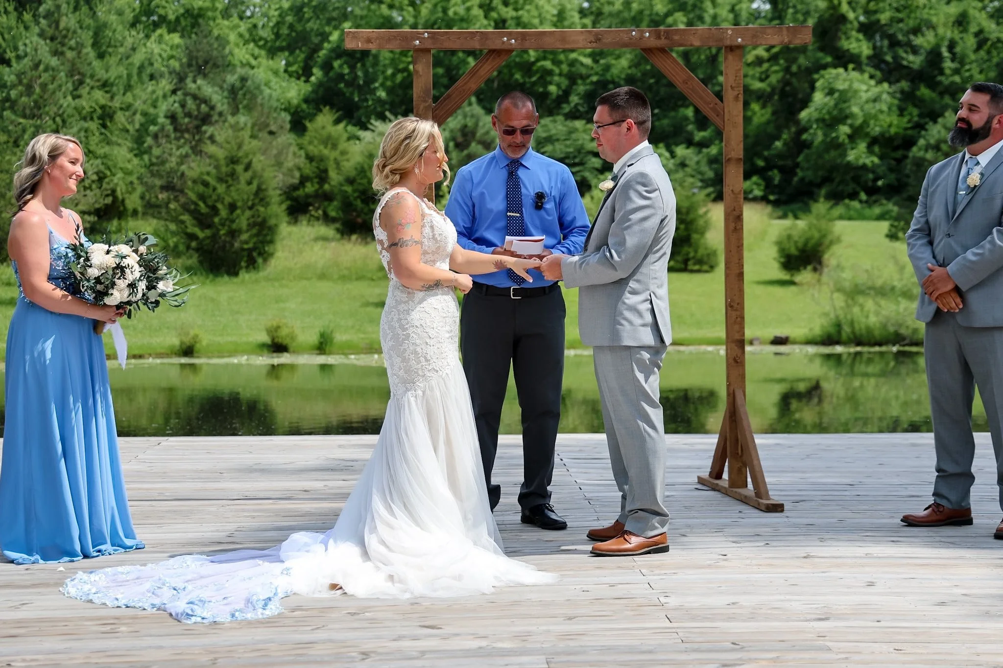 A couple getting married outdoors near a pond, exchanging vows under a simple wooden arch with an officiant, with bridesmaids and groomsmen standing nearby.