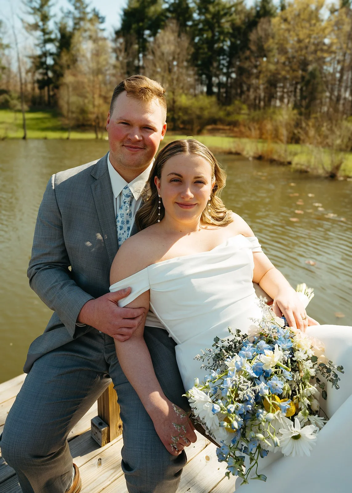 A couple on a wooden dock by a lake during daytime. The woman is wearing a white off-shoulder wedding dress and holding a bouquet of white and blue flowers. The man is in a gray suit and tie. They are smiling and posing for a photo with trees and wat