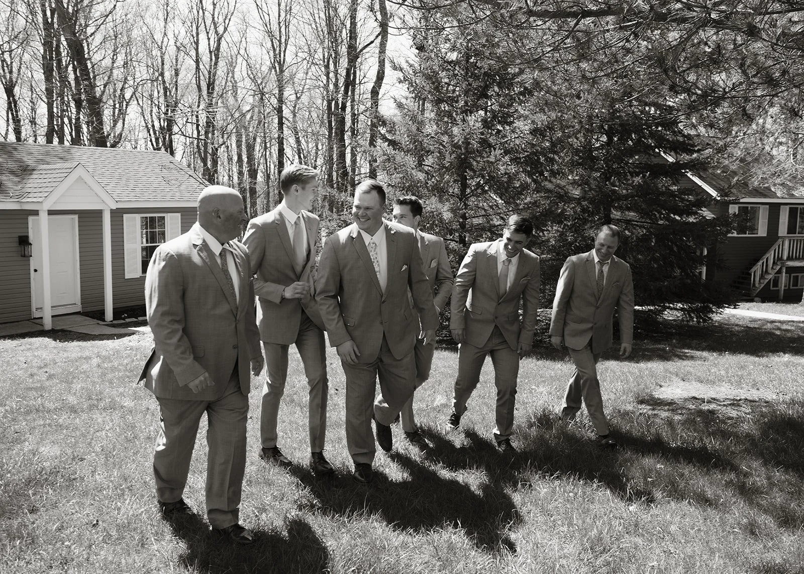 A group of six men dressed in suits walking outdoors on a sunny day, with houses and trees in the background.