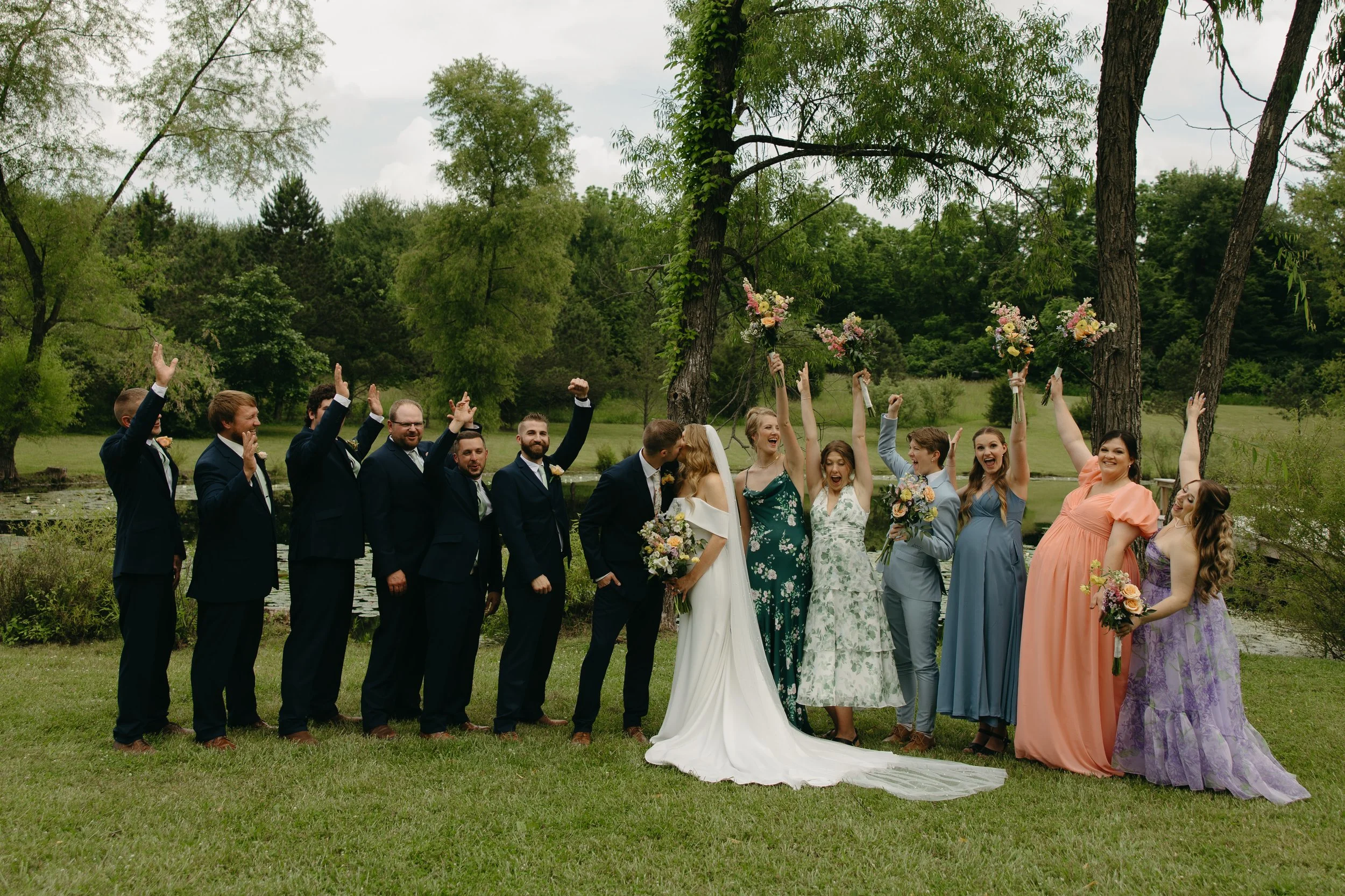 Group of wedding party members outdoors on grass, celebrating with raised hands and smiles, with trees and a pond in the background.