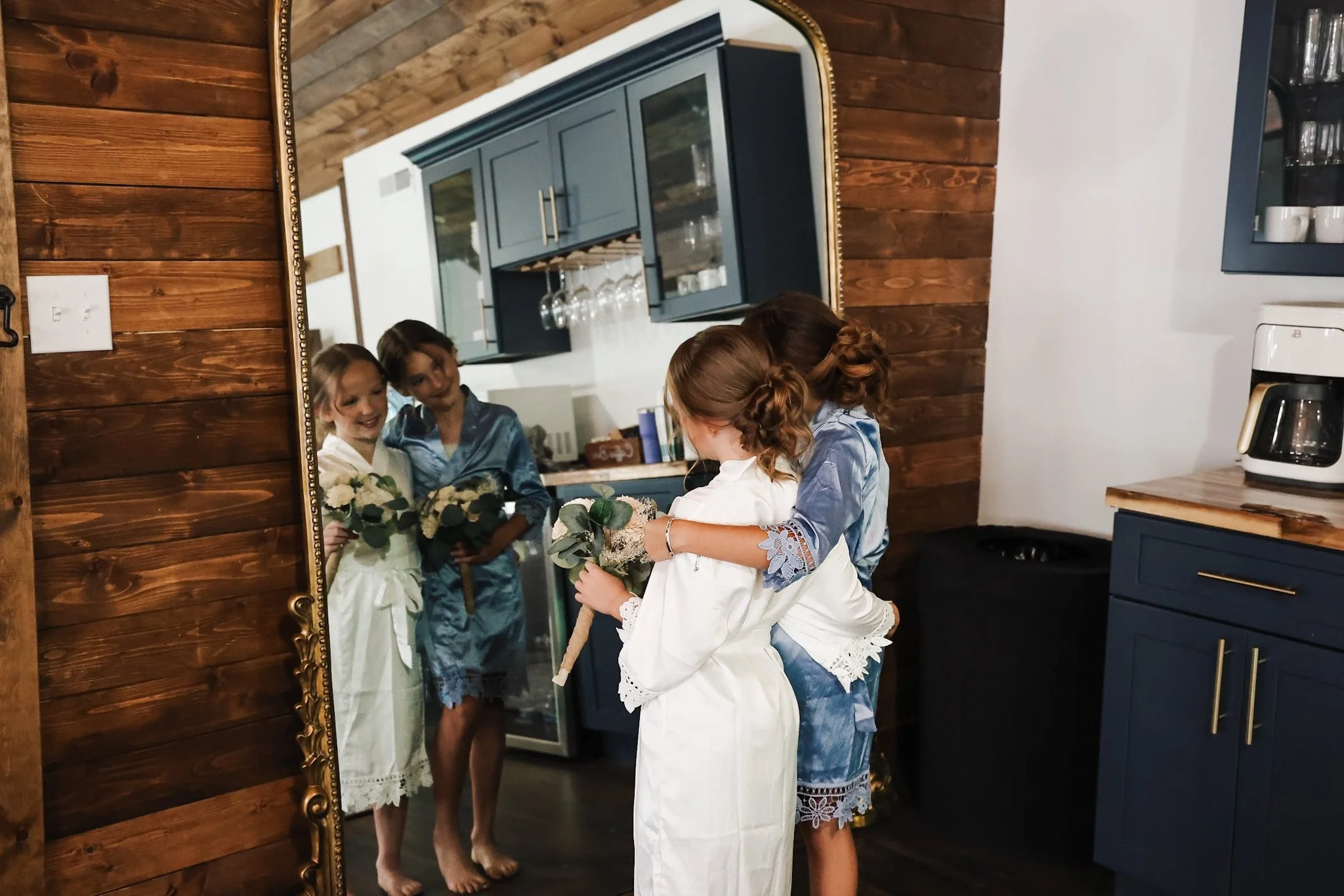 Three young girls in white and blue robes holding bouquets of flowers, standing barefoot in front of a mirror in a cozy kitchen with blue cabinets and wooden walls, looking at their reflections.
