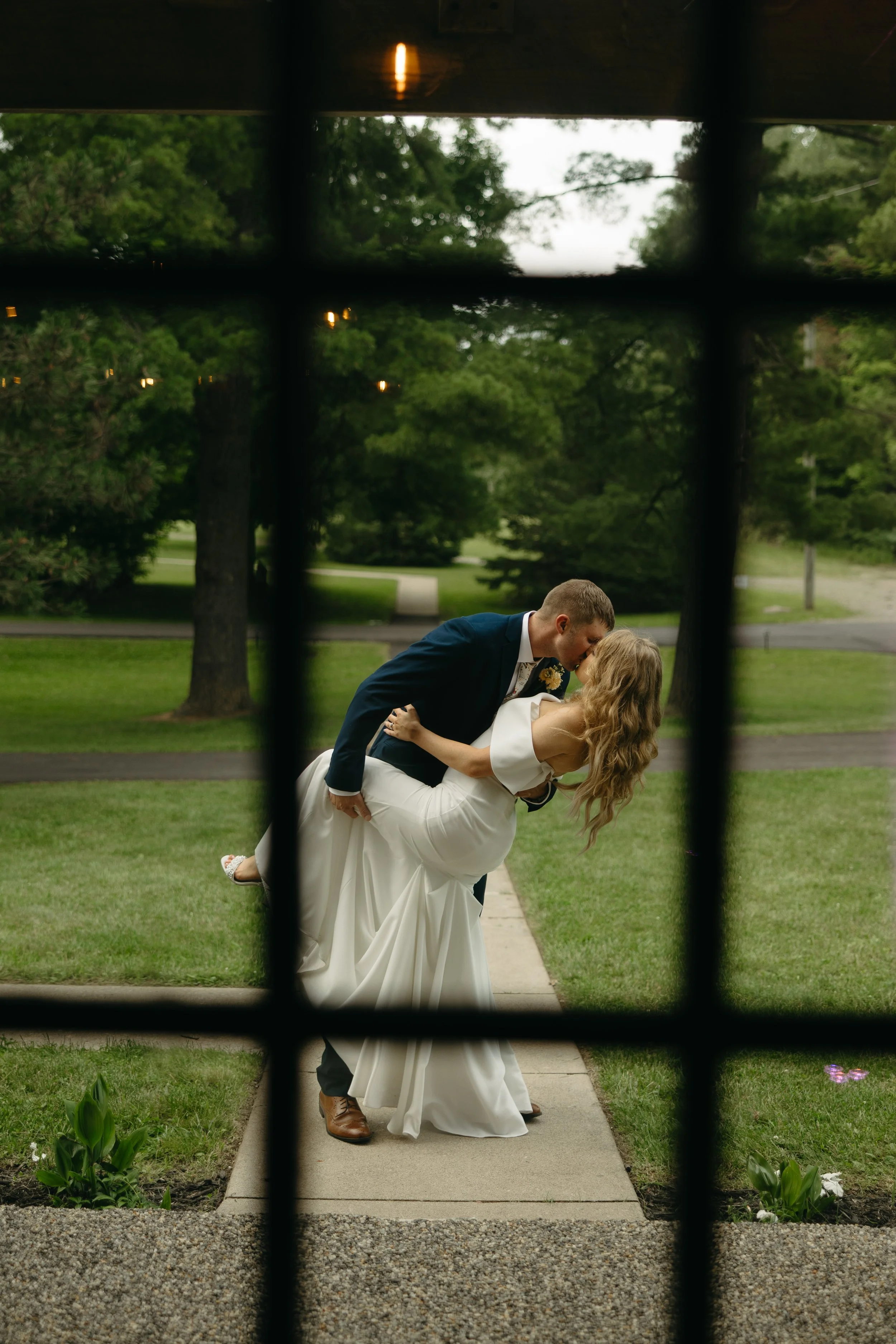 A newlywed couple sharing a kiss outdoors on a grass pathway, viewed through a window with metal bars.