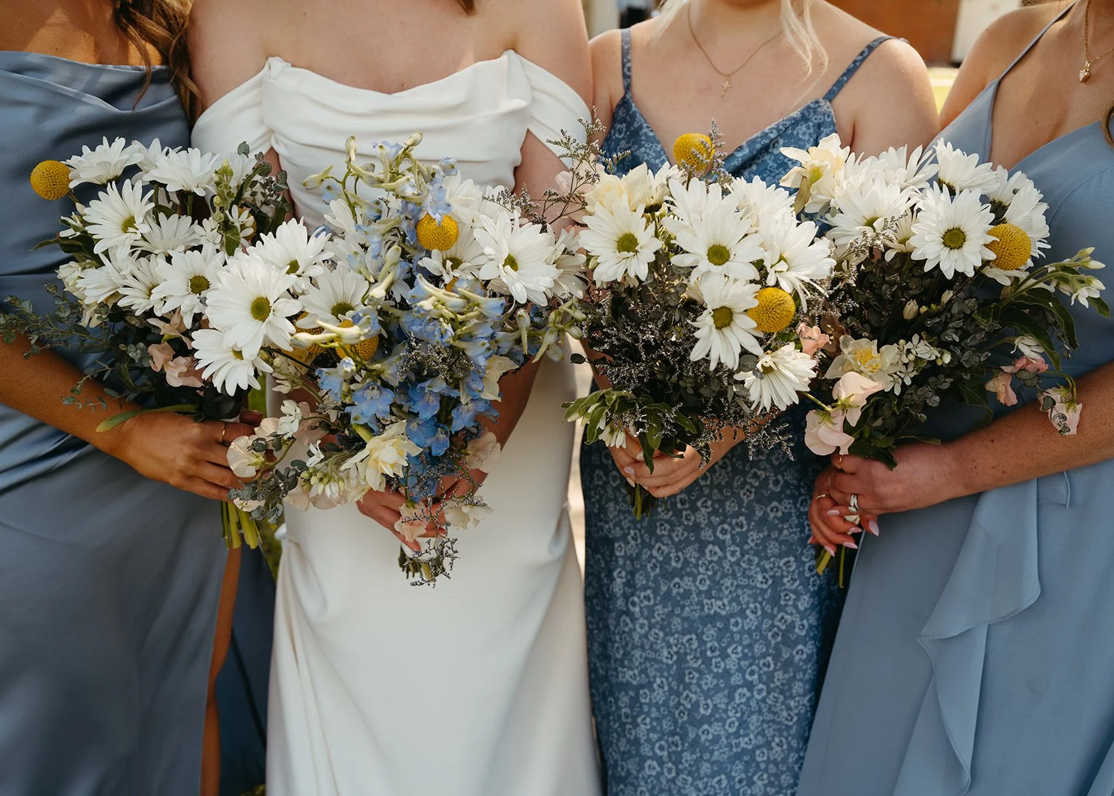 Close-up of women holding bouquets of white daisies and blue flowers, from the waist up at a wedding or special event.
