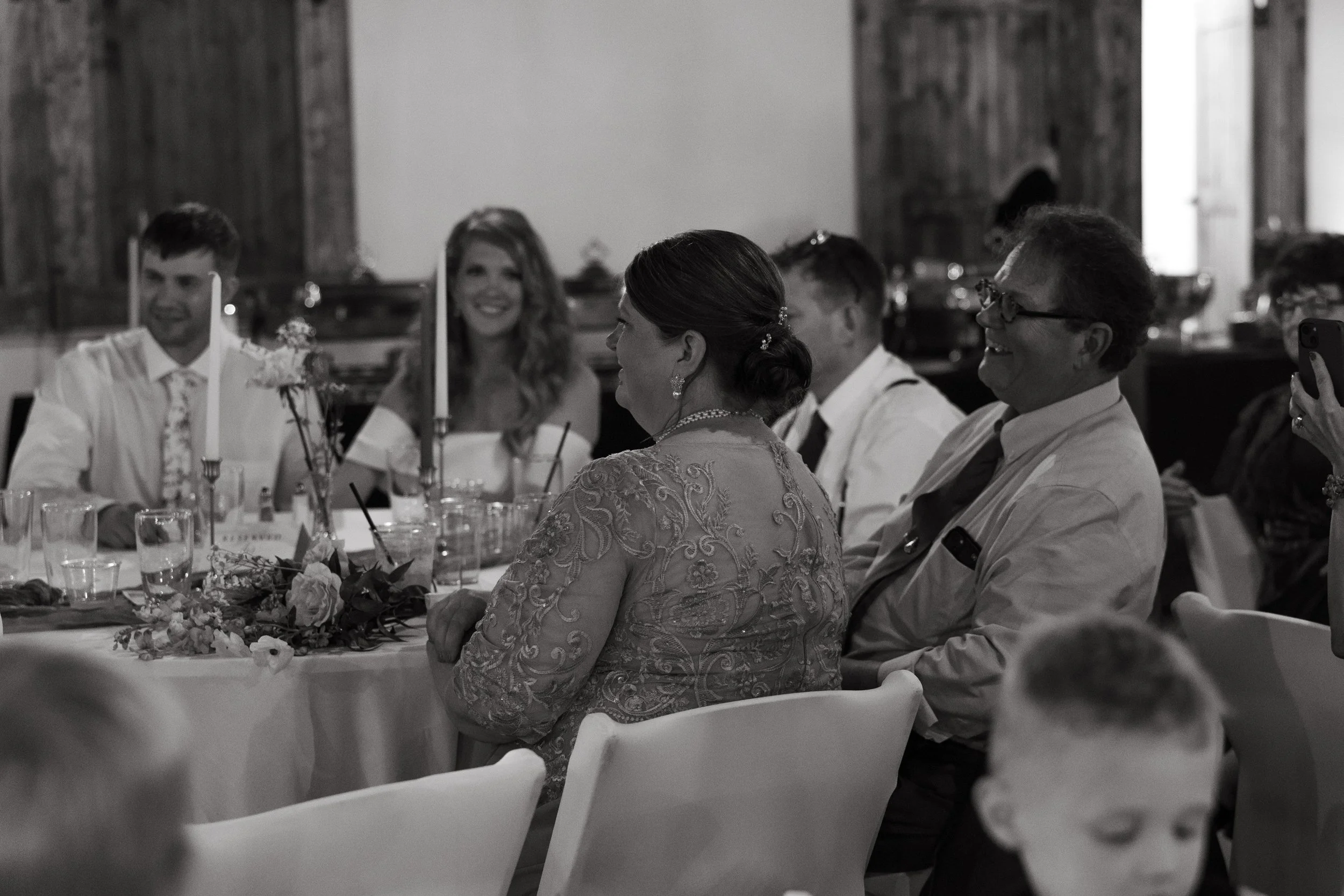 People seated at a dinner table during a celebration, smiling and engaging with each other, with candles and floral arrangements on the table, in a rustic indoor setting.