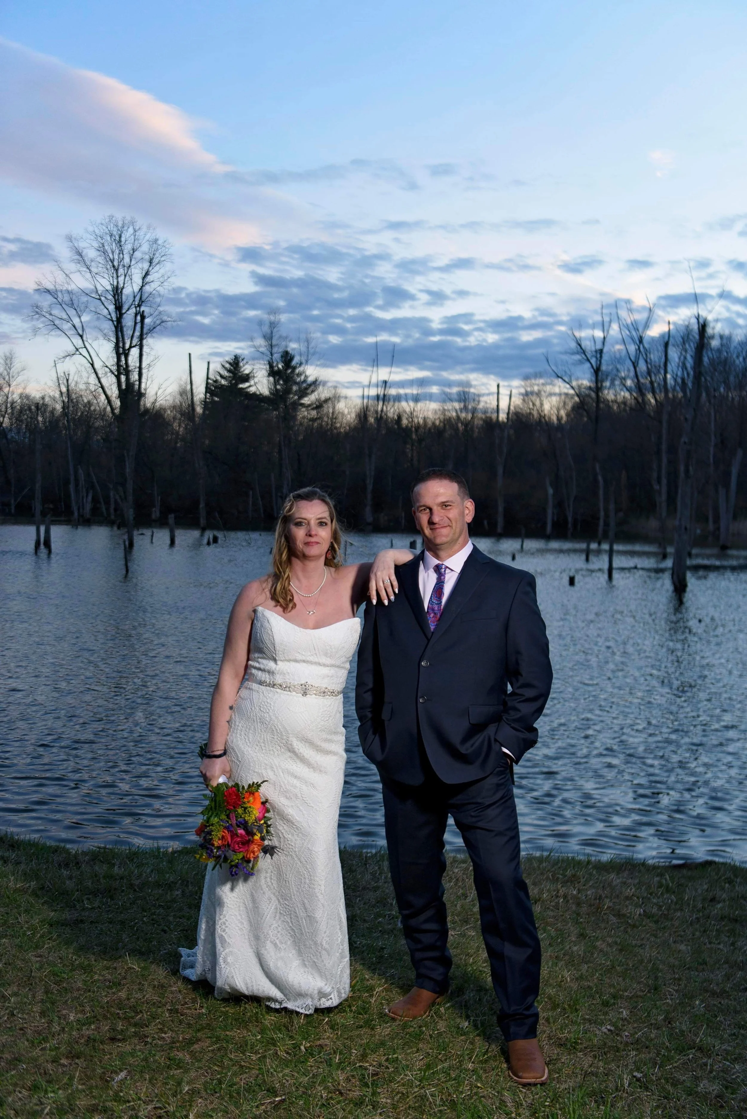 A bride and groom stand together near a body of water during sunset or dusk, with leafless trees and a partly cloudy sky in the background. The bride wears a strapless white wedding dress and holds a colorful bouquet, while the groom wears a dark sui