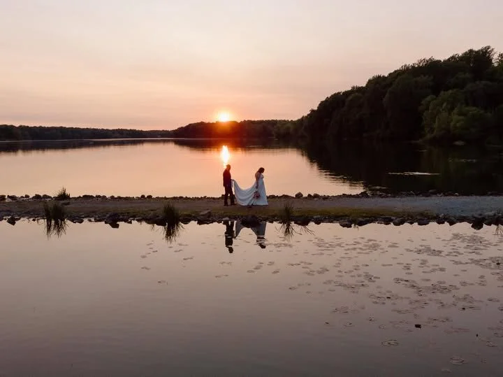 A couple standing on a narrow strip of land by a calm river at sunset, holding hands with the sun setting behind them and trees on the opposite riverbank.