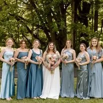 A bride and six bridesmaids standing outdoors in a wooded area, all holding bouquets and dressed in pastel-colored dresses.