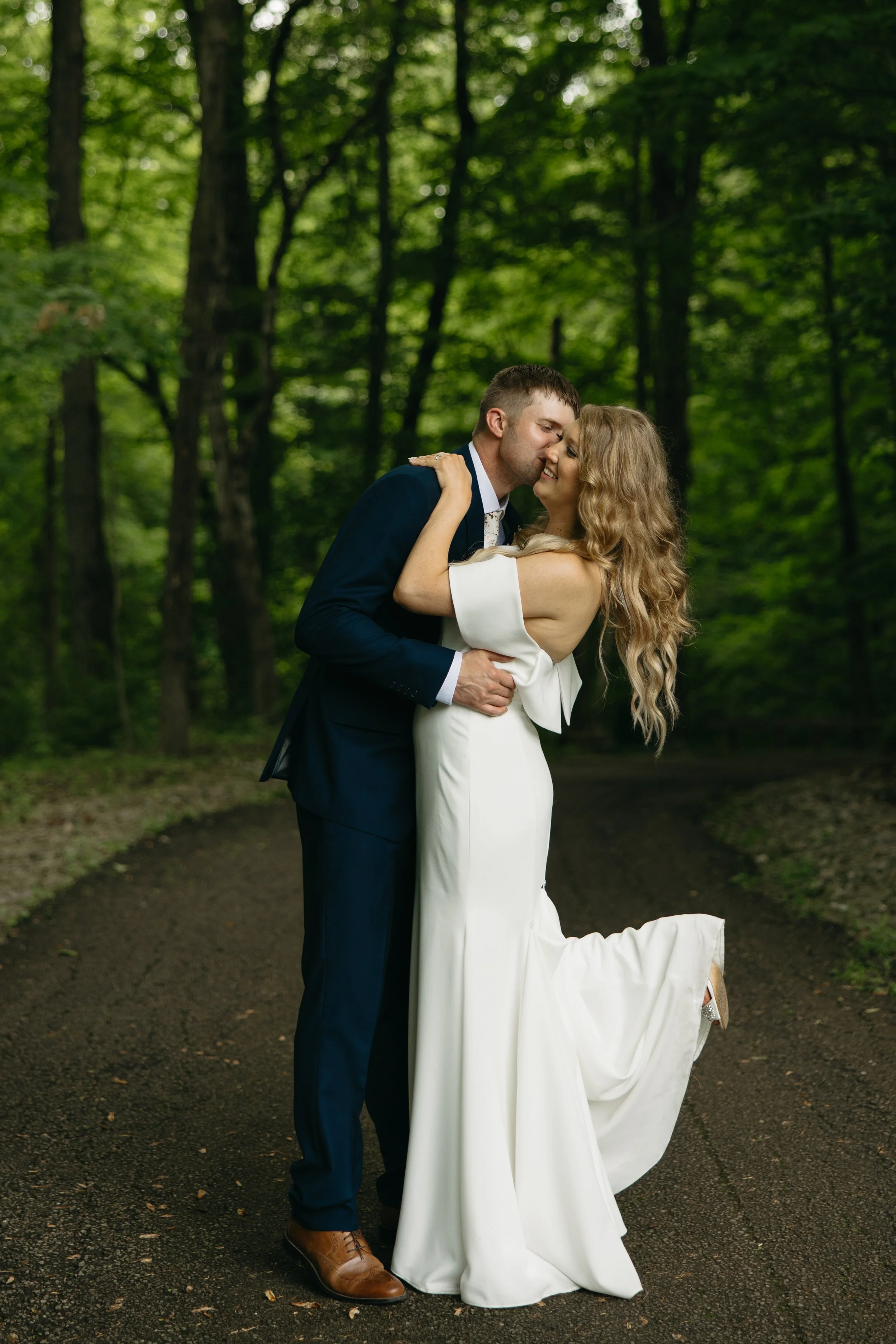 A newlywed couple kissing and smiling in a forested area, with the groom wearing a dark suit and the bride in a white wedding dress.