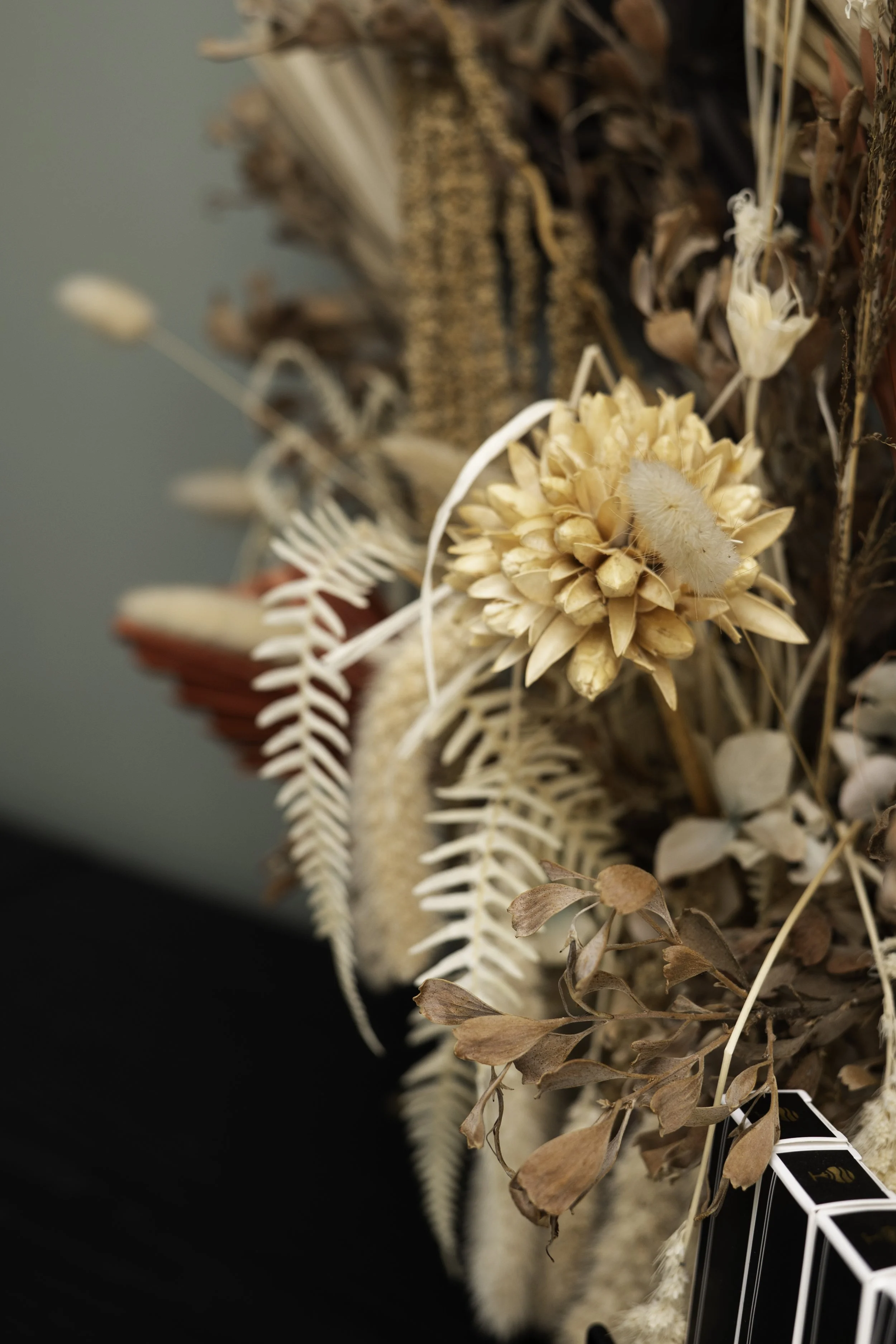 A close-up of a display of artificial dried flowers and foliage, including a large beige flower with a fluffy center, brown leaves, and white fern-like leaves, arranged on a black background.