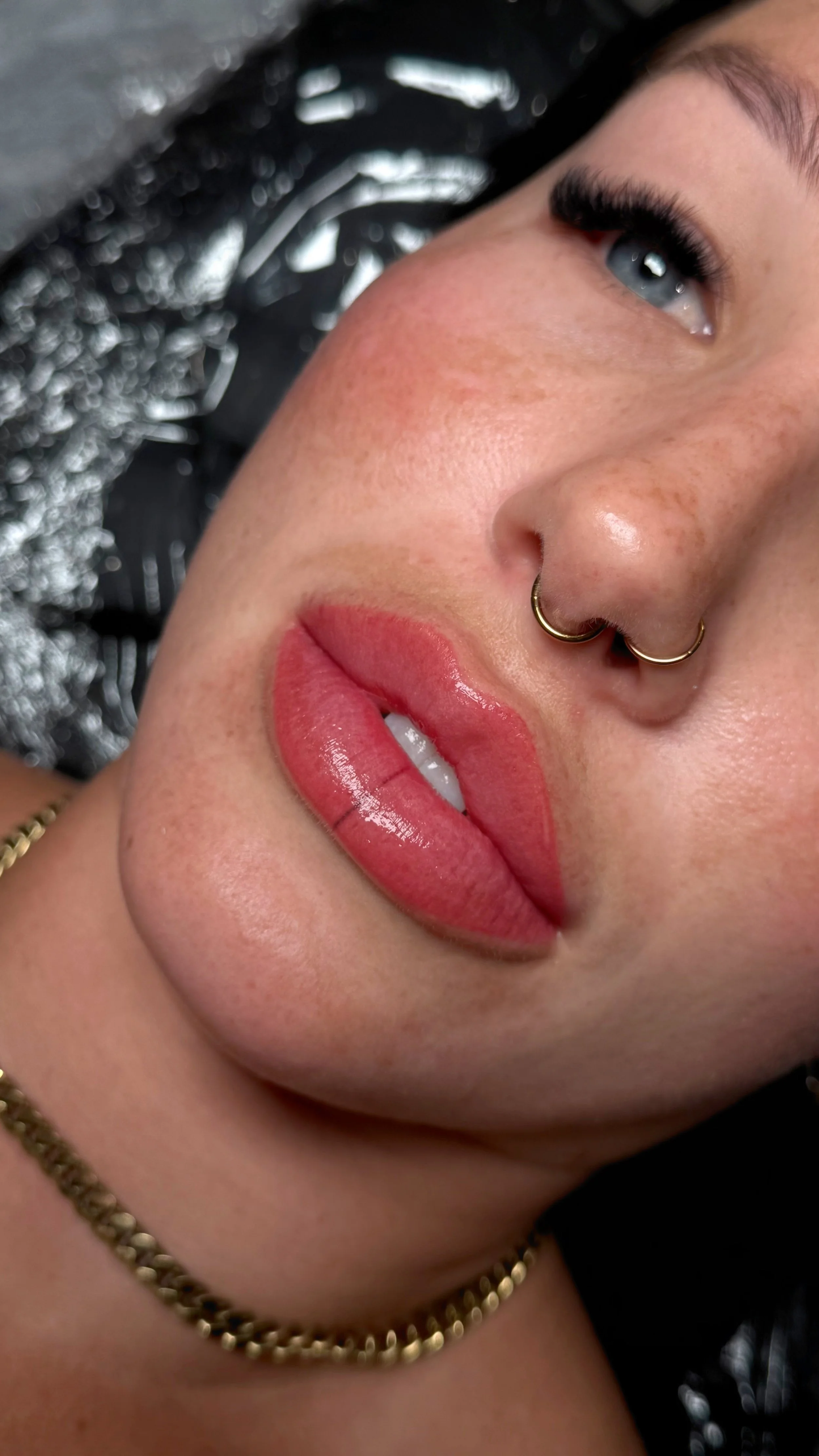 Close-up of a person's face showing their eye, nose with a septum piercing, full lips with glossy lipstick, freckles, and a gold chain necklace.