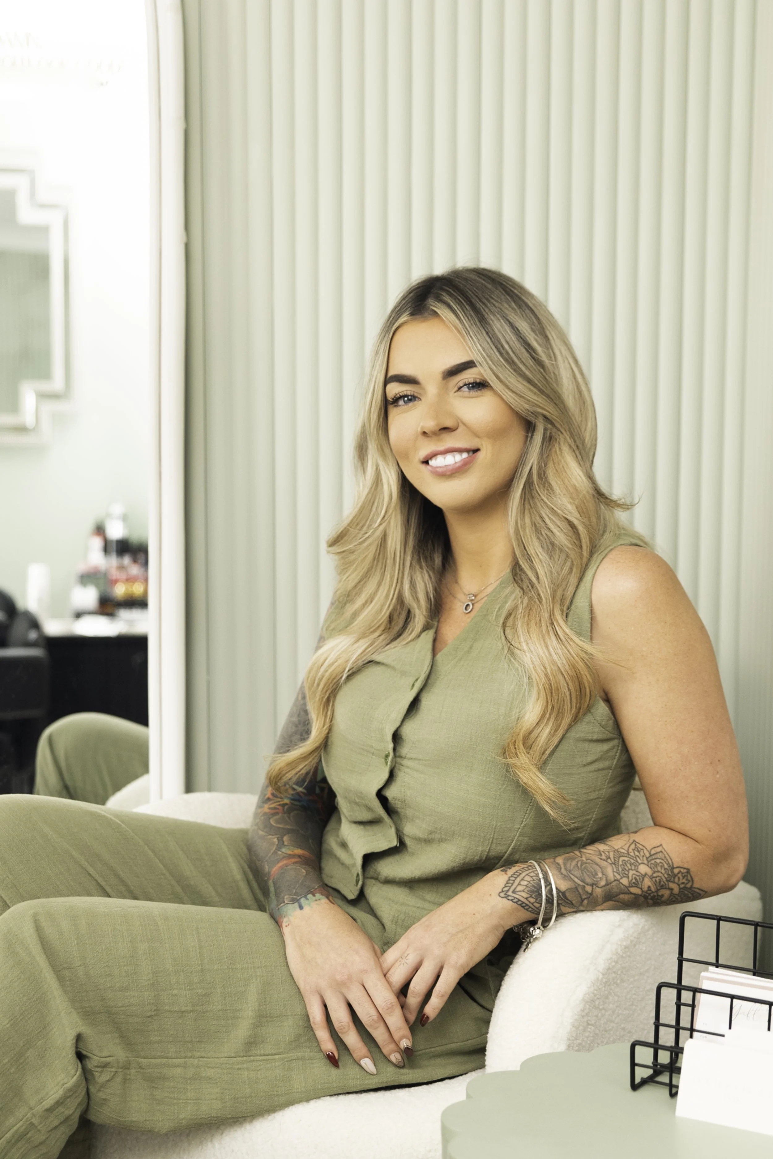 A woman with long, wavy blonde hair sitting on a green chair in a salon or studio, smiling at the camera. She has tattoos on her arms and wears a sleeveless olive-green top.