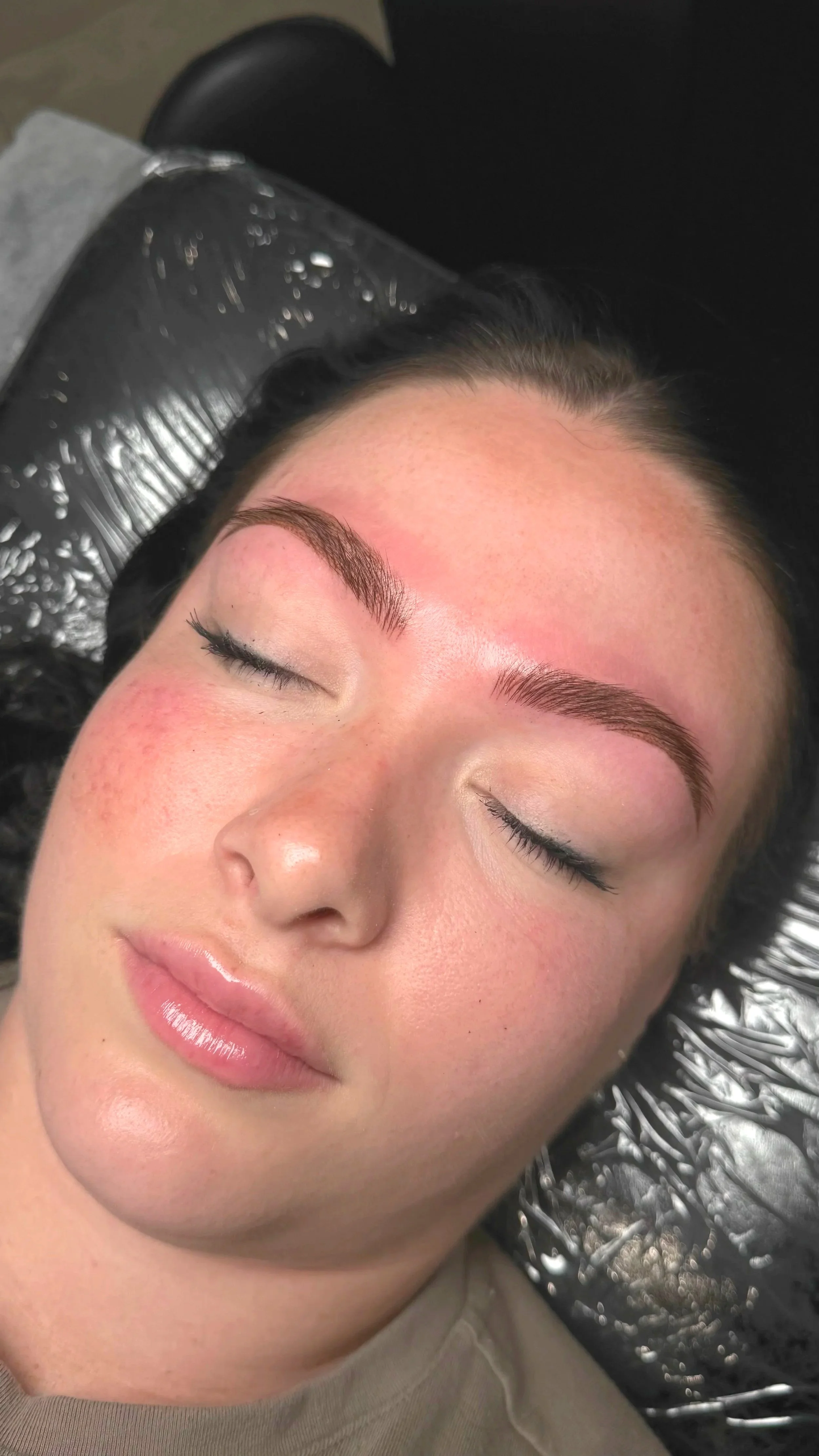 Woman with freshly done eyebrow microblading, lying down with eyes closed, on a black protective mat.