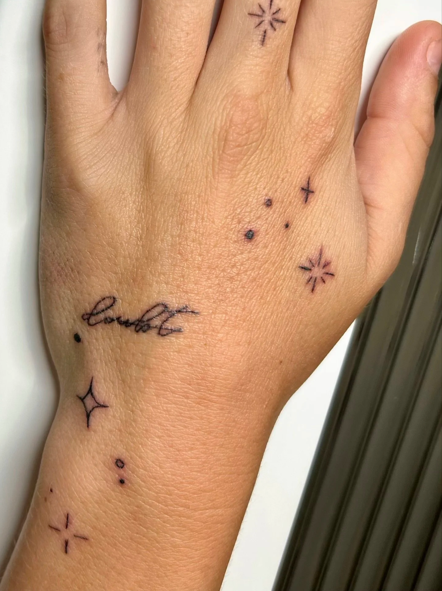 Close-up of a person's hand with various small black ink tattoos, including stars, dots, and cursive writing on the fingers and wrist.